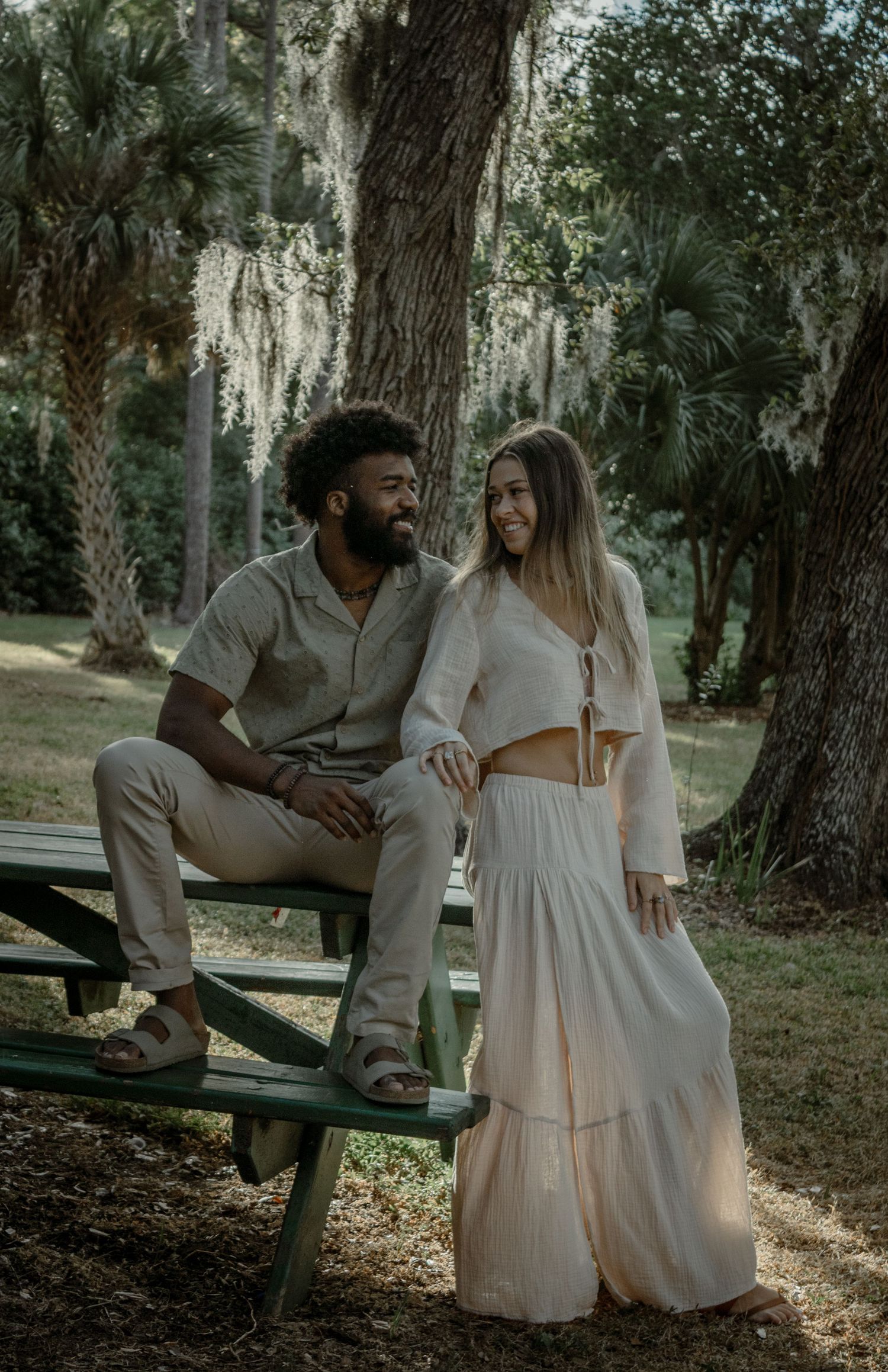 A couple sits together on a picnic table surrounded by Spanish moss and palm trees in a romantic outdoor setting.