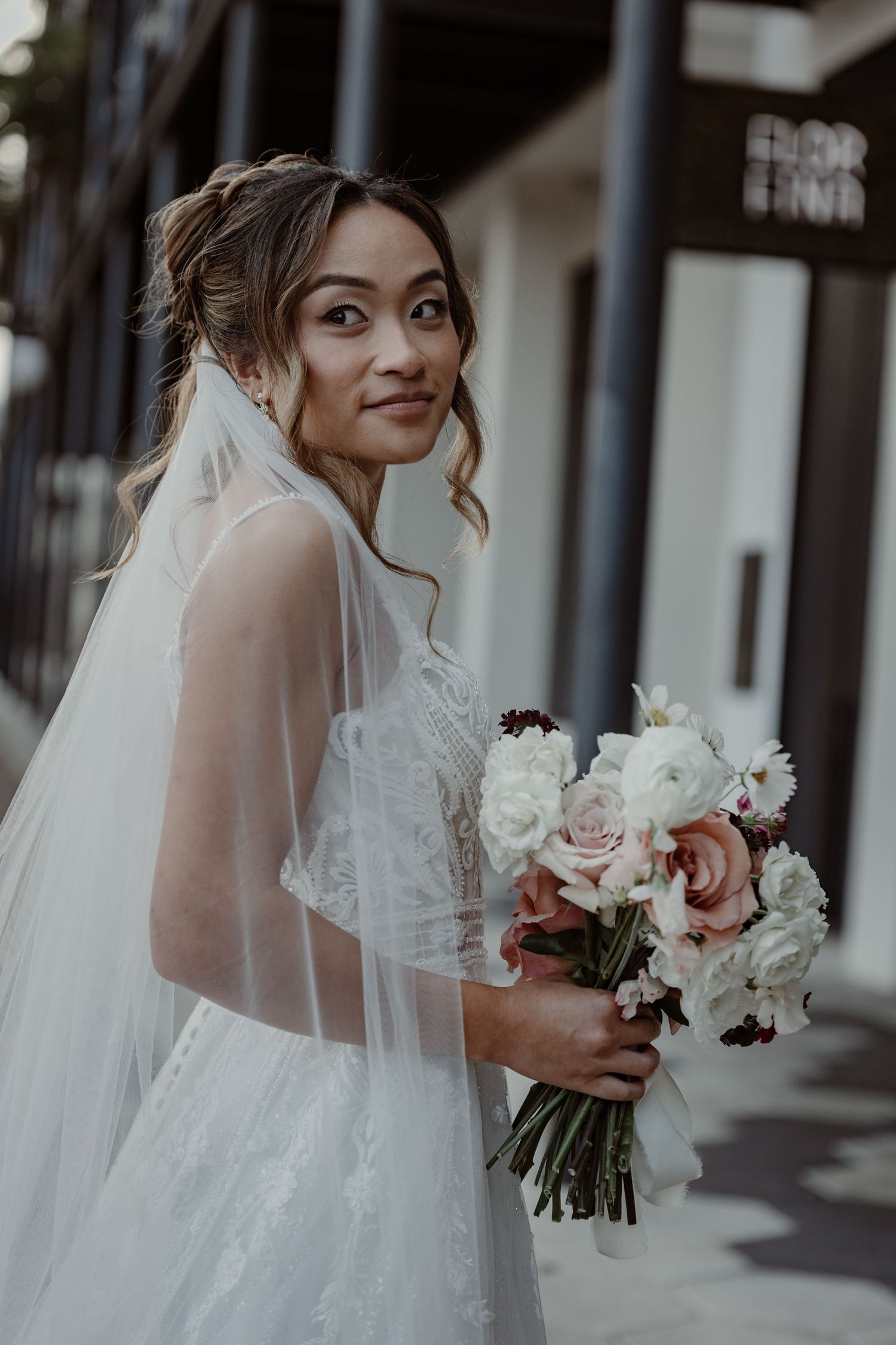 A bride in a flowing white dress and veil smiles over her shoulder while holding a pink and white bouquet.