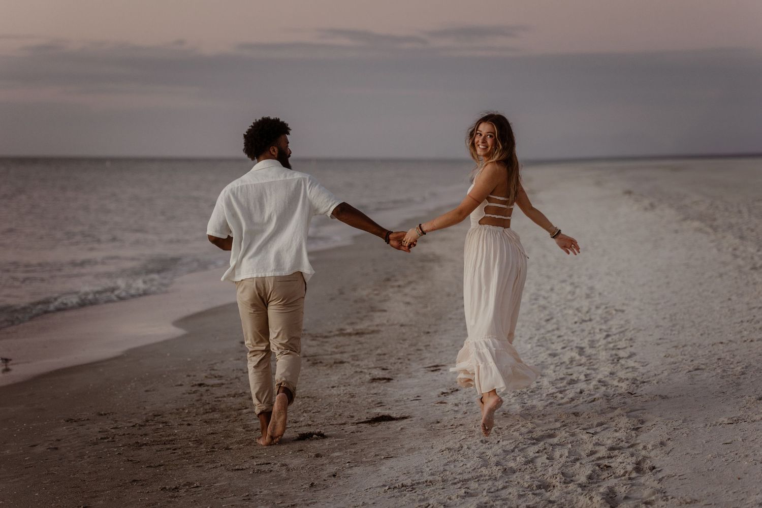 Couple in white outfits walking hand in hand along a misty beach at sunset.