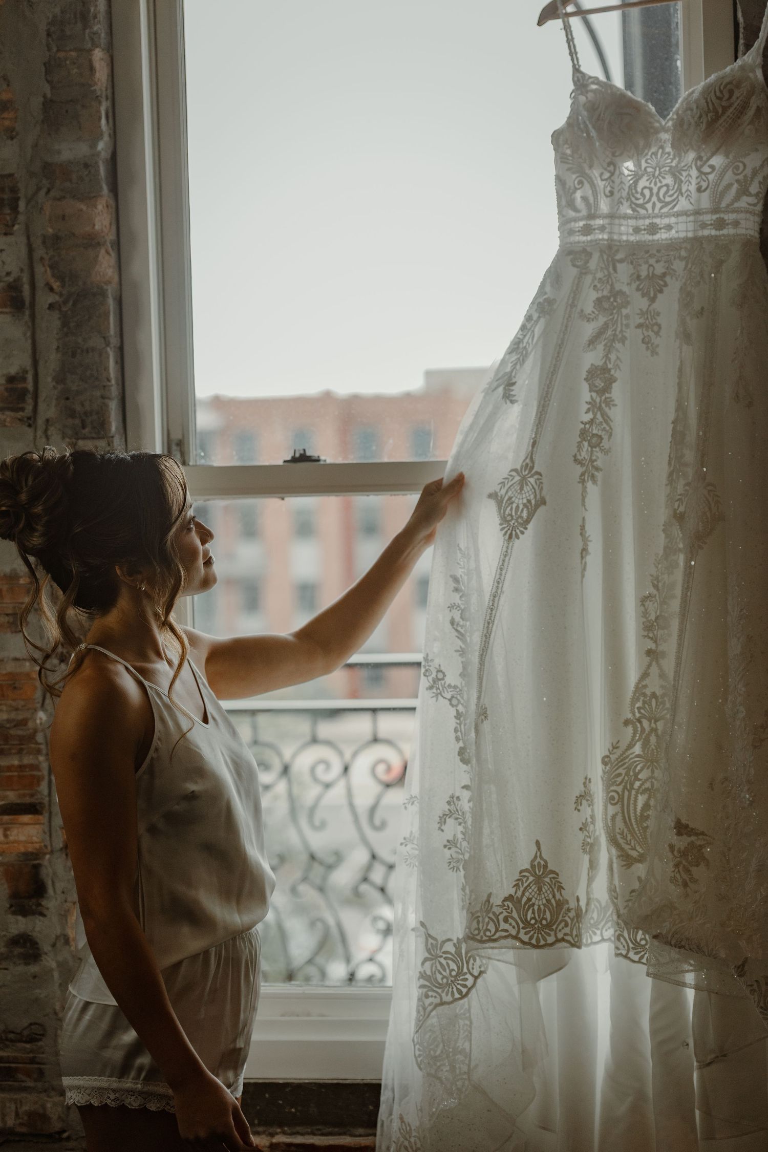 A person in a white dress stands by a window with delicate lace curtains in a rustic brick interior.