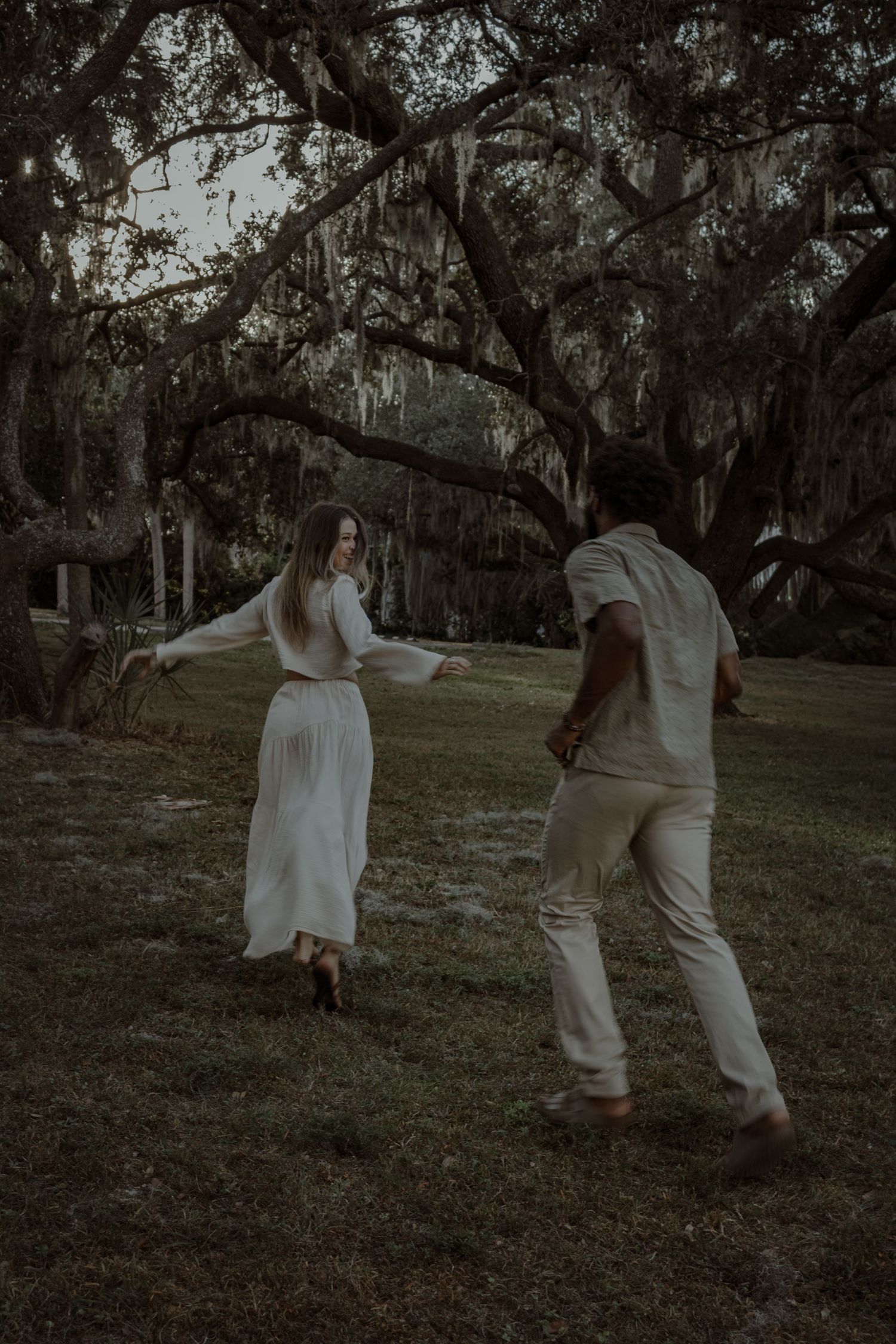 A couple in white clothing dances playfully together in a park setting with Spanish moss hanging from trees.