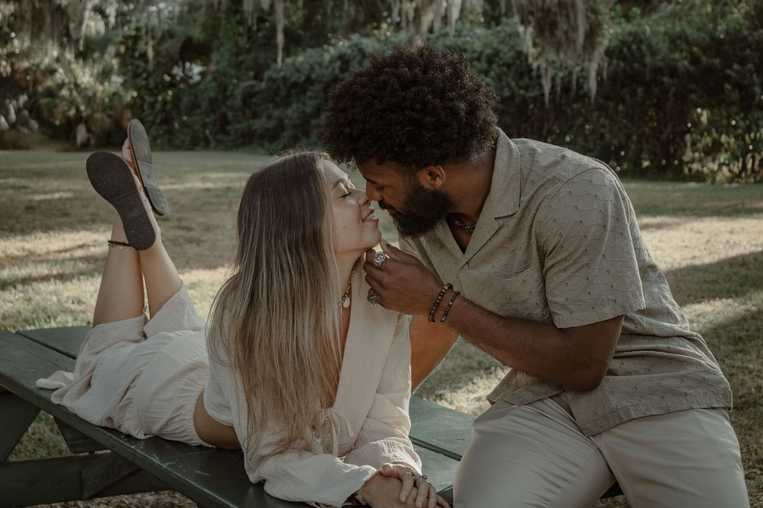 A romantic couple shares intimate moments together in a natural outdoor setting with sunlit trees in the background.