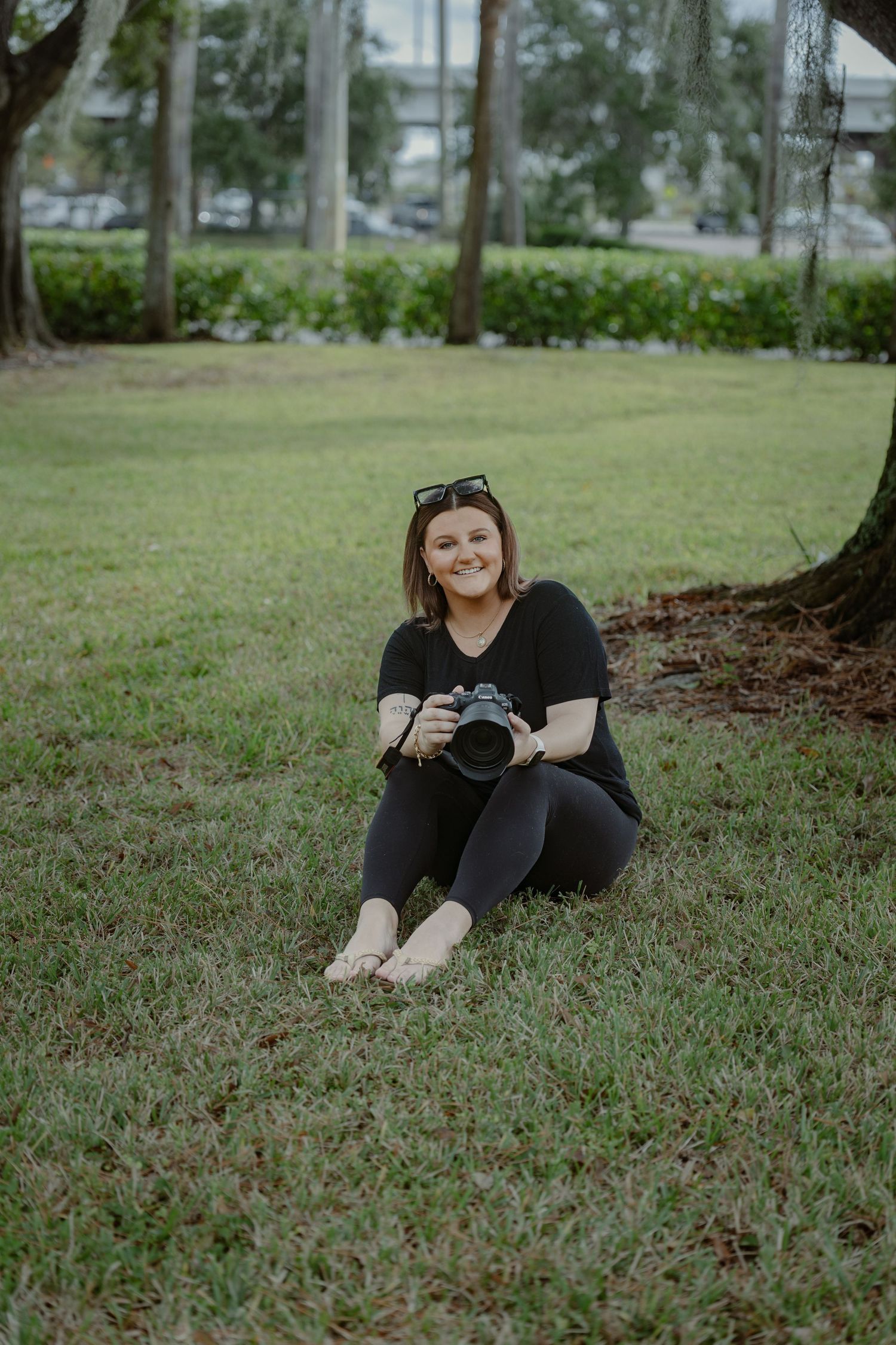 Photographer sits on grass holding camera in outdoor setting with trees and greenery in background.