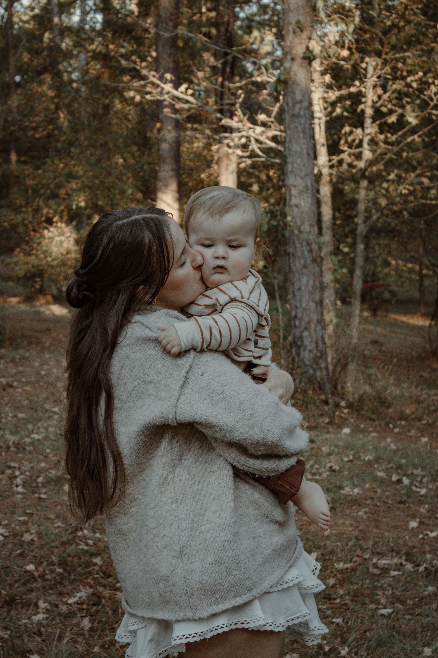 Mother in gray sweater embraces and kisses baby in white dress during autumn forest photoshoot.