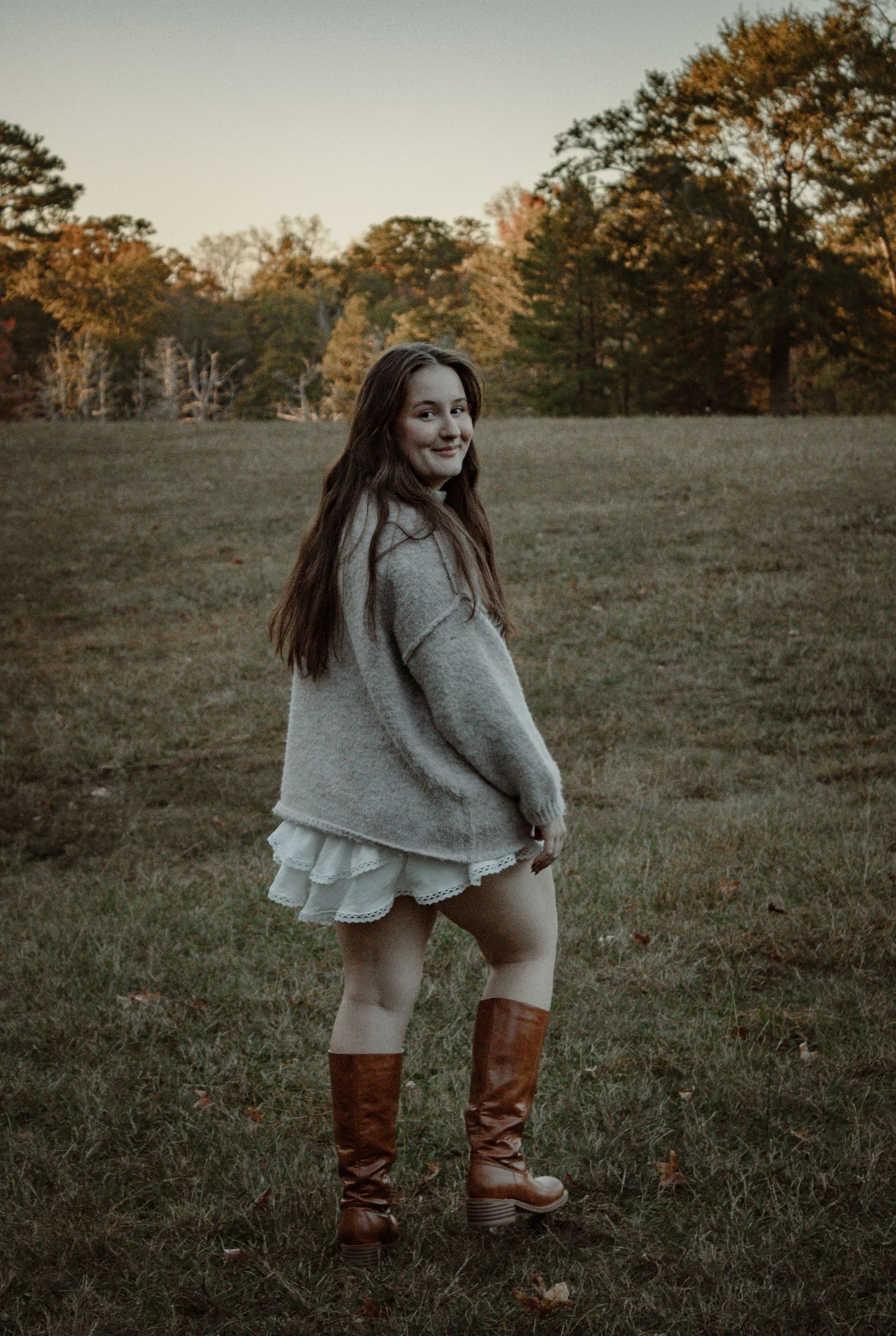 A person in a cozy gray sweater and brown boots poses in a field during golden hour at sunset.