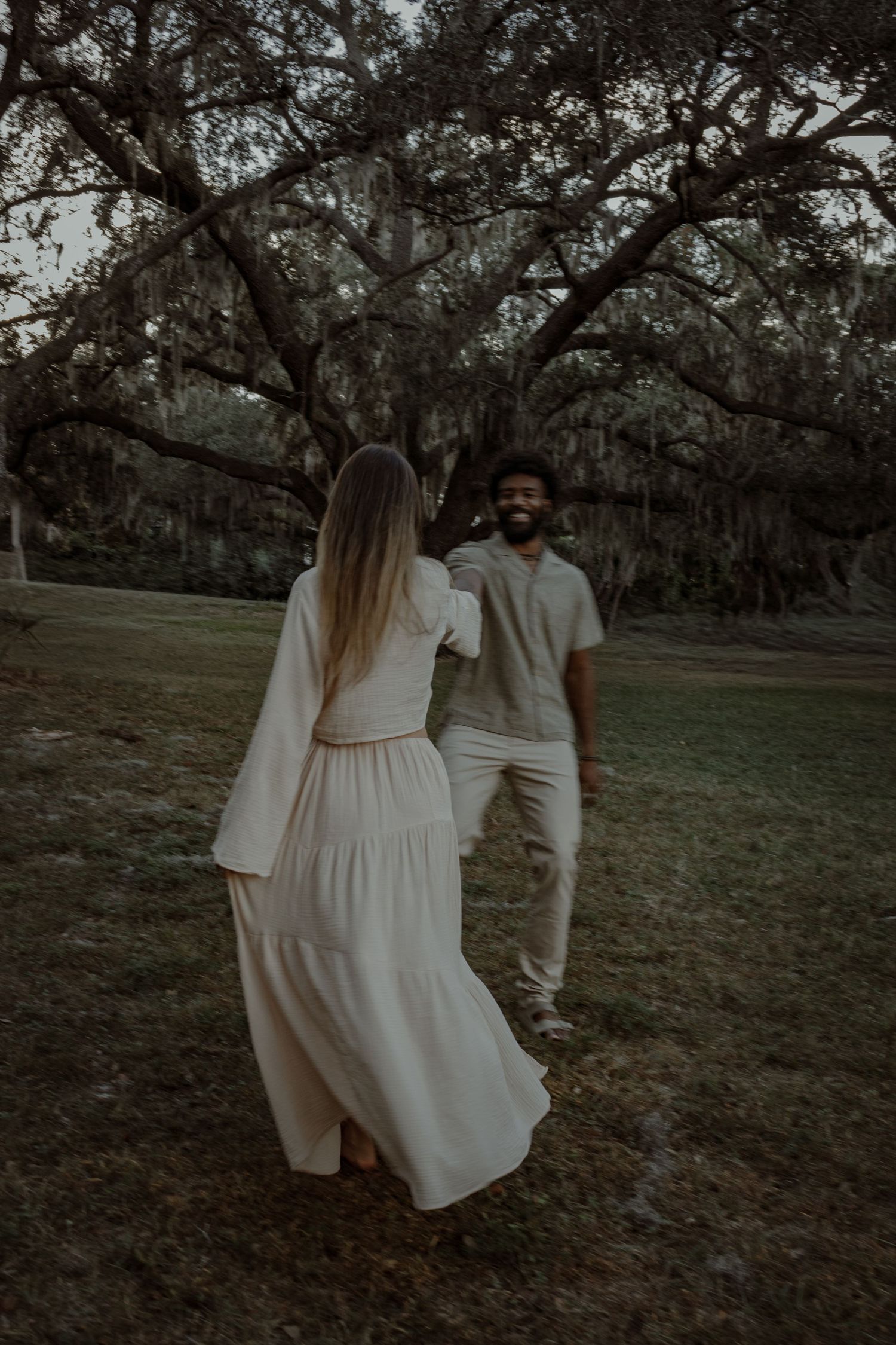 A couple in white outfits dances playfully in a park with moss-covered trees in the background.