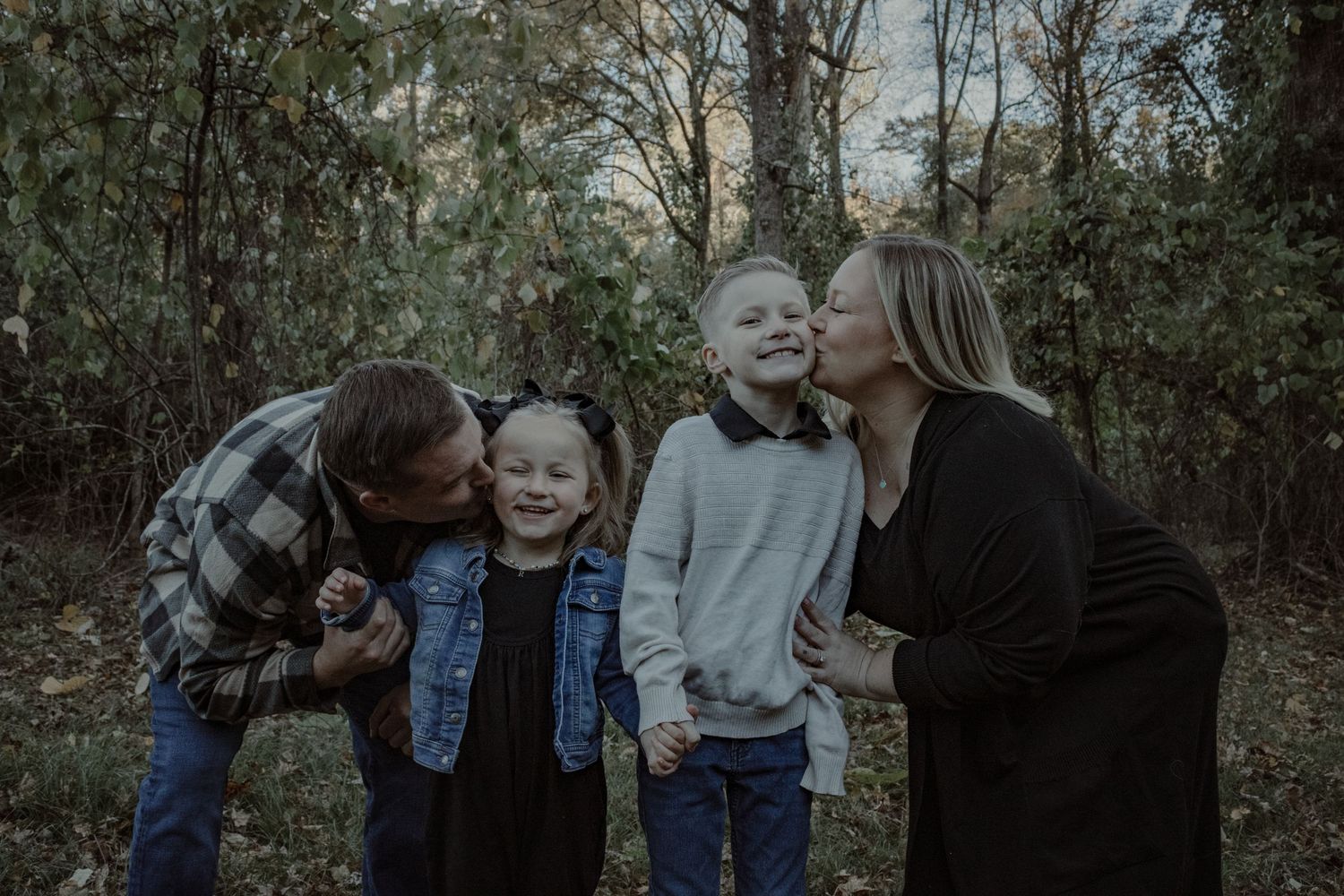 A family group poses together in a wooded outdoor setting during autumn wearing coordinated casual attire.
