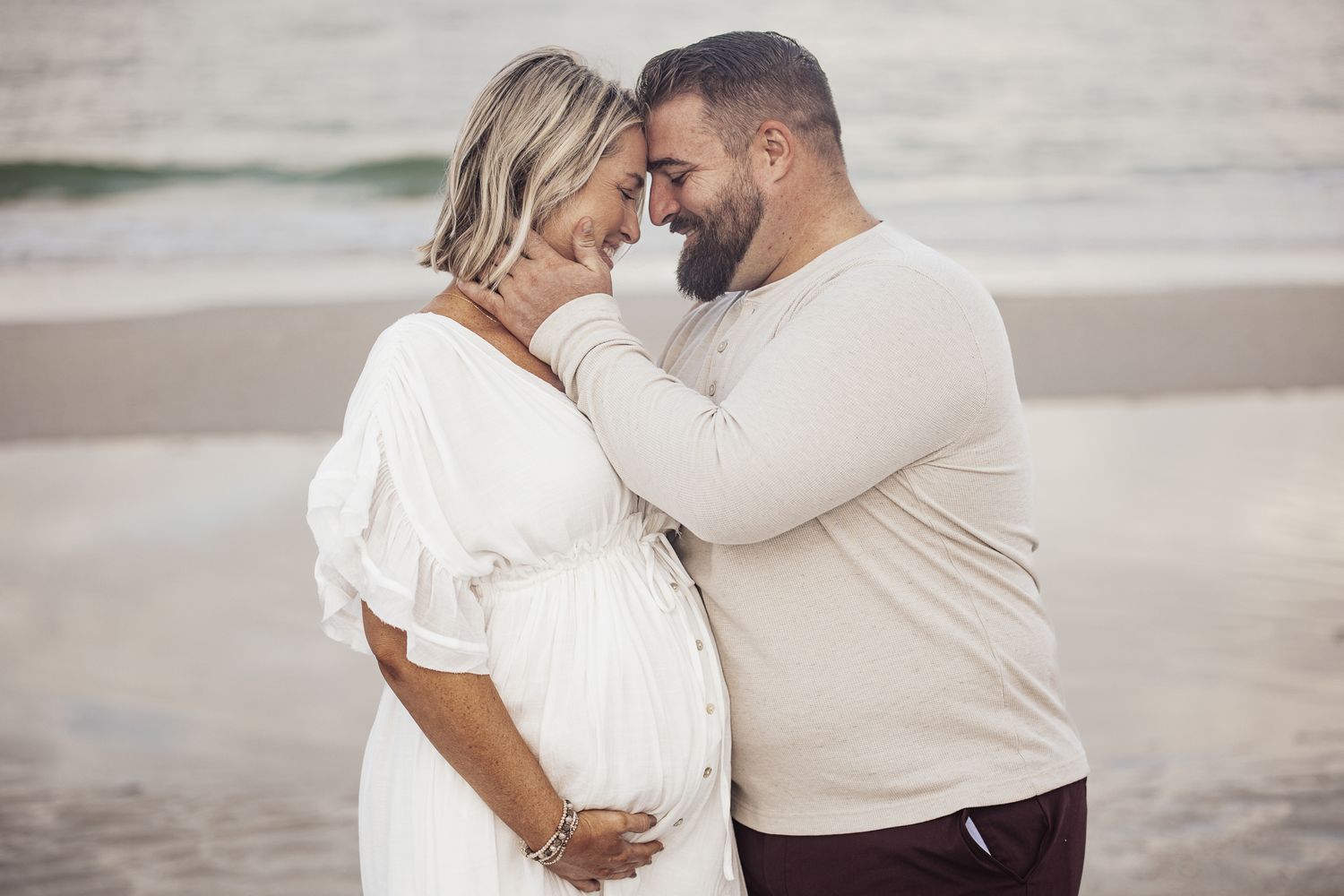 A romantic beach photoshoot captures tender moments between a couple in white outfits against a misty coastal backdrop.
