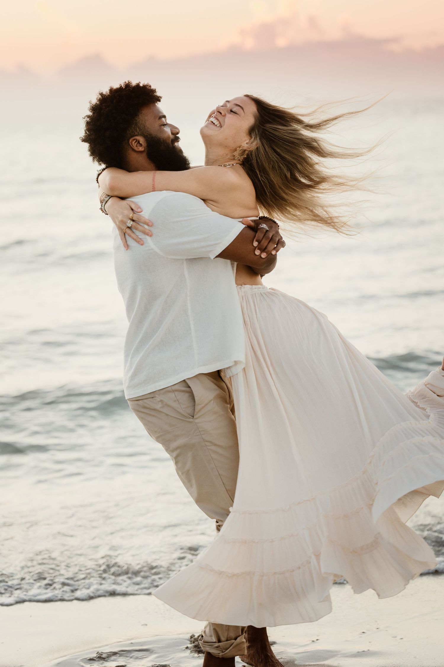 A joyful couple embraces and spins on the beach during sunset with waves in the background.