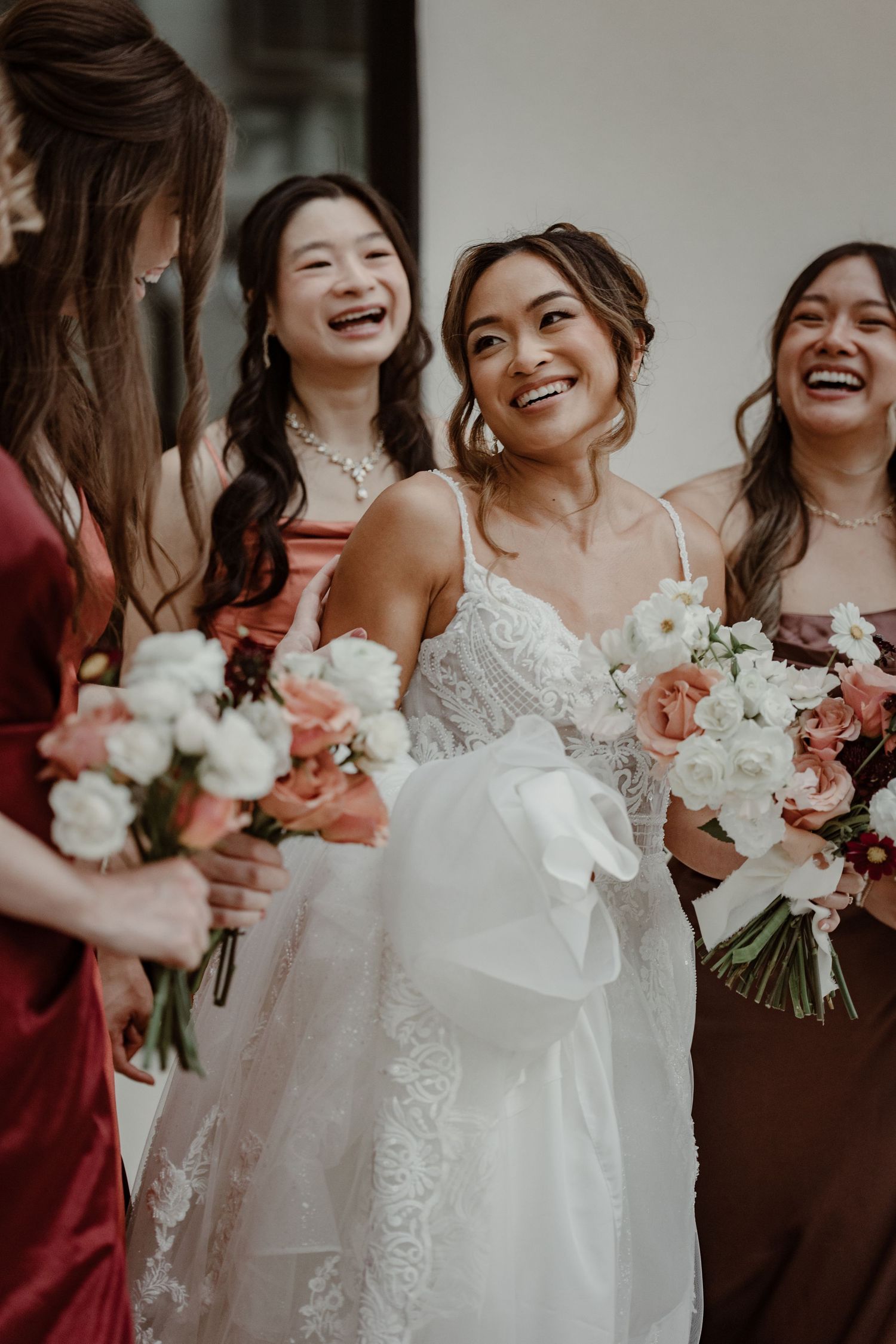 Wedding party members in burgundy dresses laugh together while holding white floral bouquets.