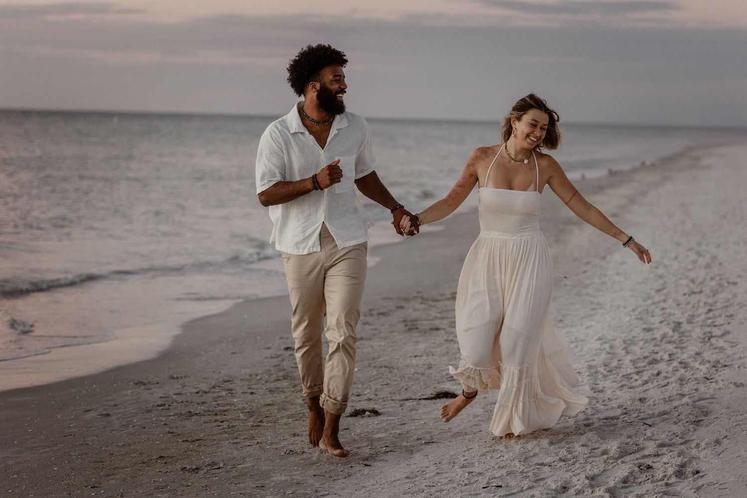 A couple in white attire walks hand in hand along a serene beach during sunset creating a romantic atmosphere.