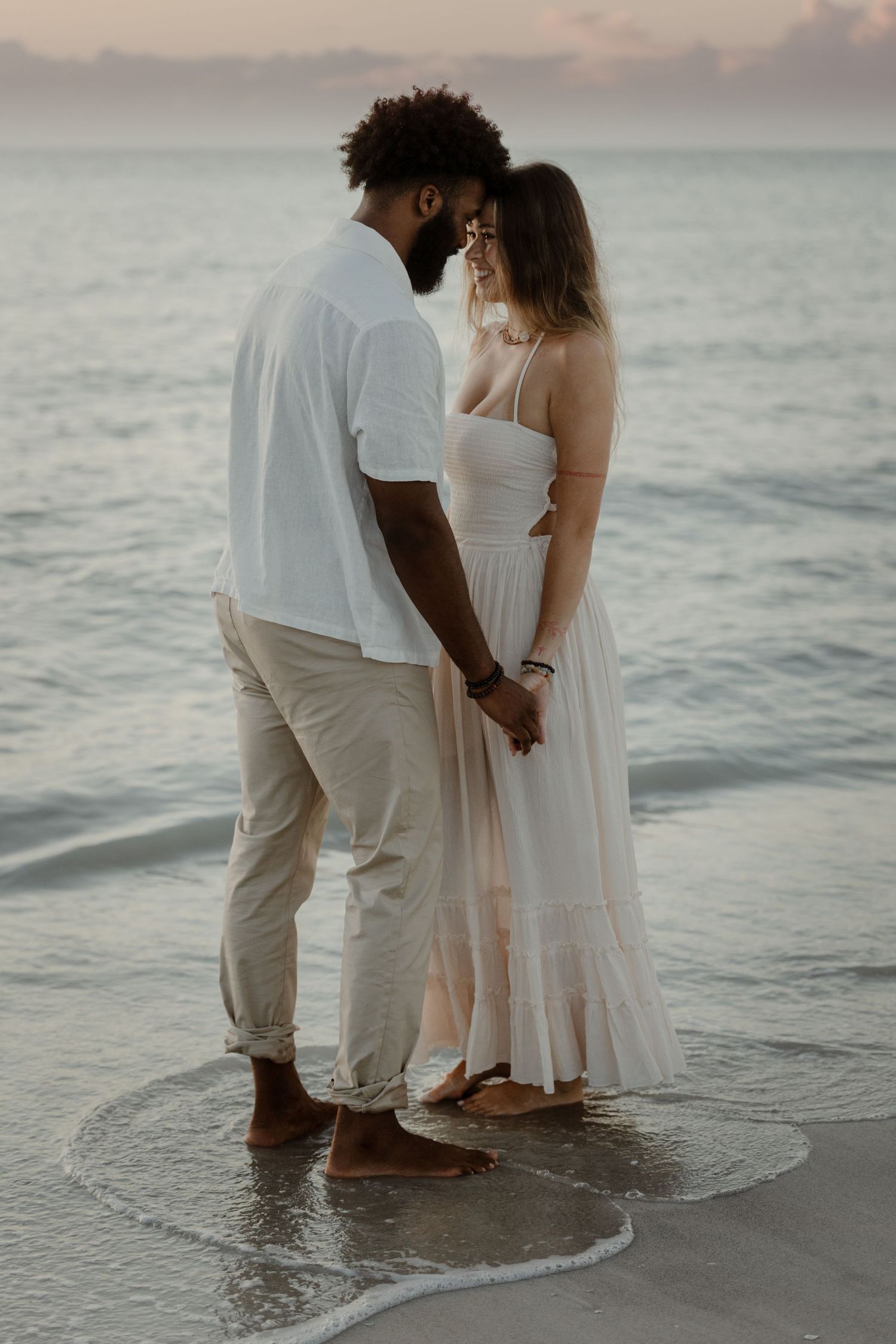 A couple stands together at the ocean's edge during sunset wearing flowing white outfits.