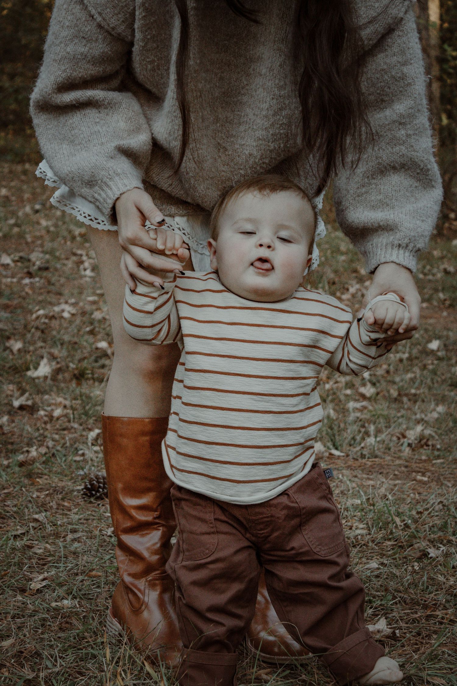 A child in a striped shirt and brown pants holds an adult's hand while walking outdoors.