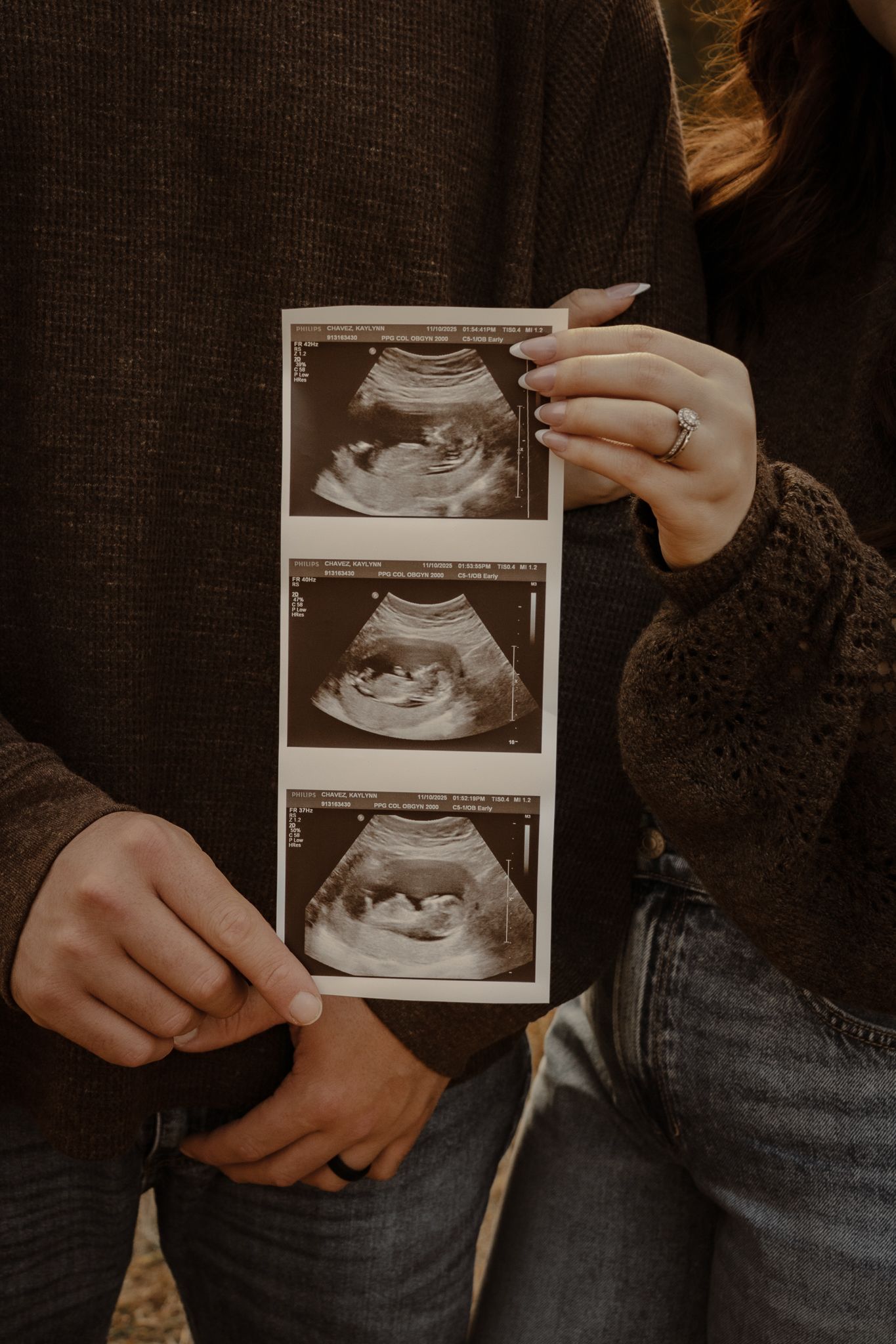 Someone wearing a dark sweater holds ultrasound photos while sitting in jeans, showing a pregnancy announcement moment.