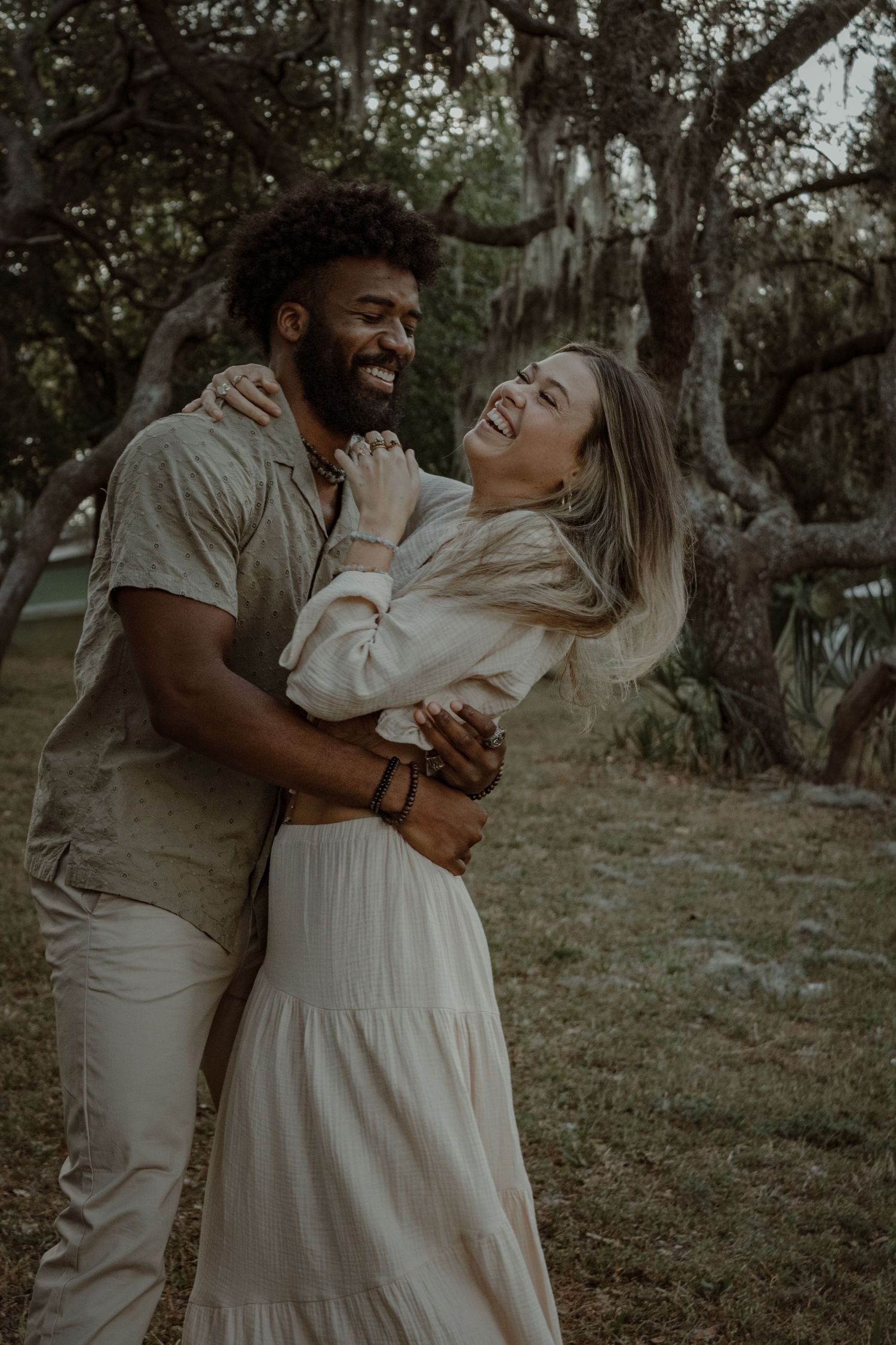 A joyful couple embraces and laughs together during a romantic outdoor photoshoot under moss-draped trees.