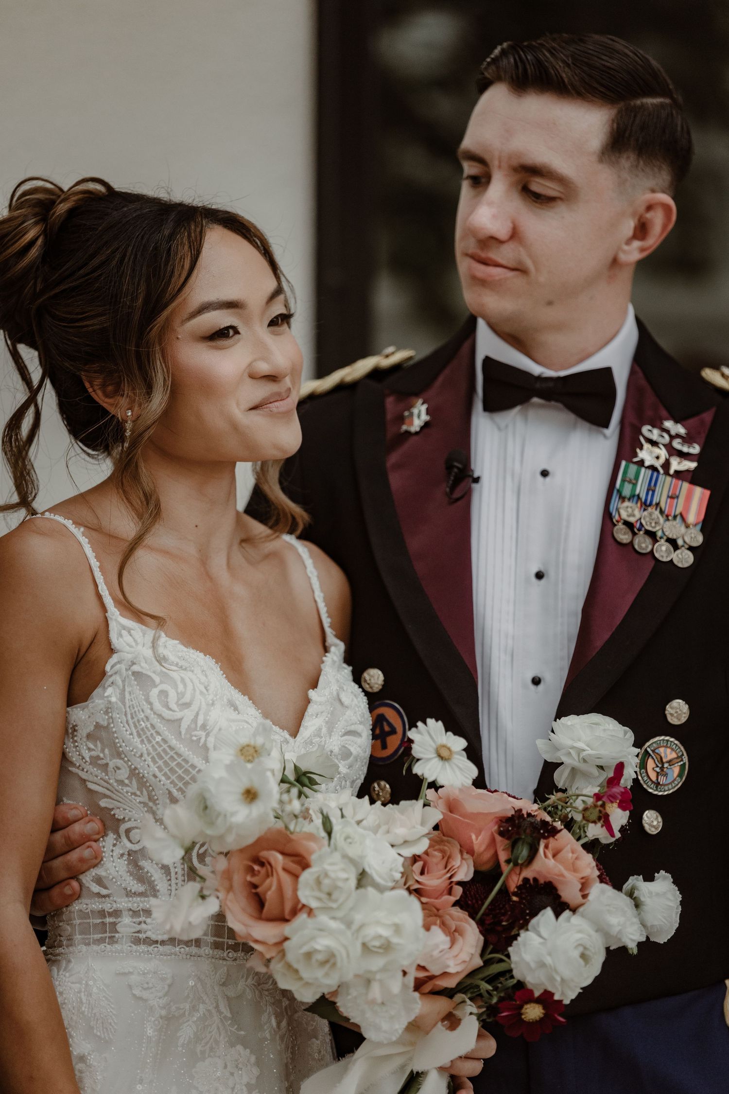 A bride in a lace wedding dress holds a white and pink floral bouquet next to a groom in military dress uniform.