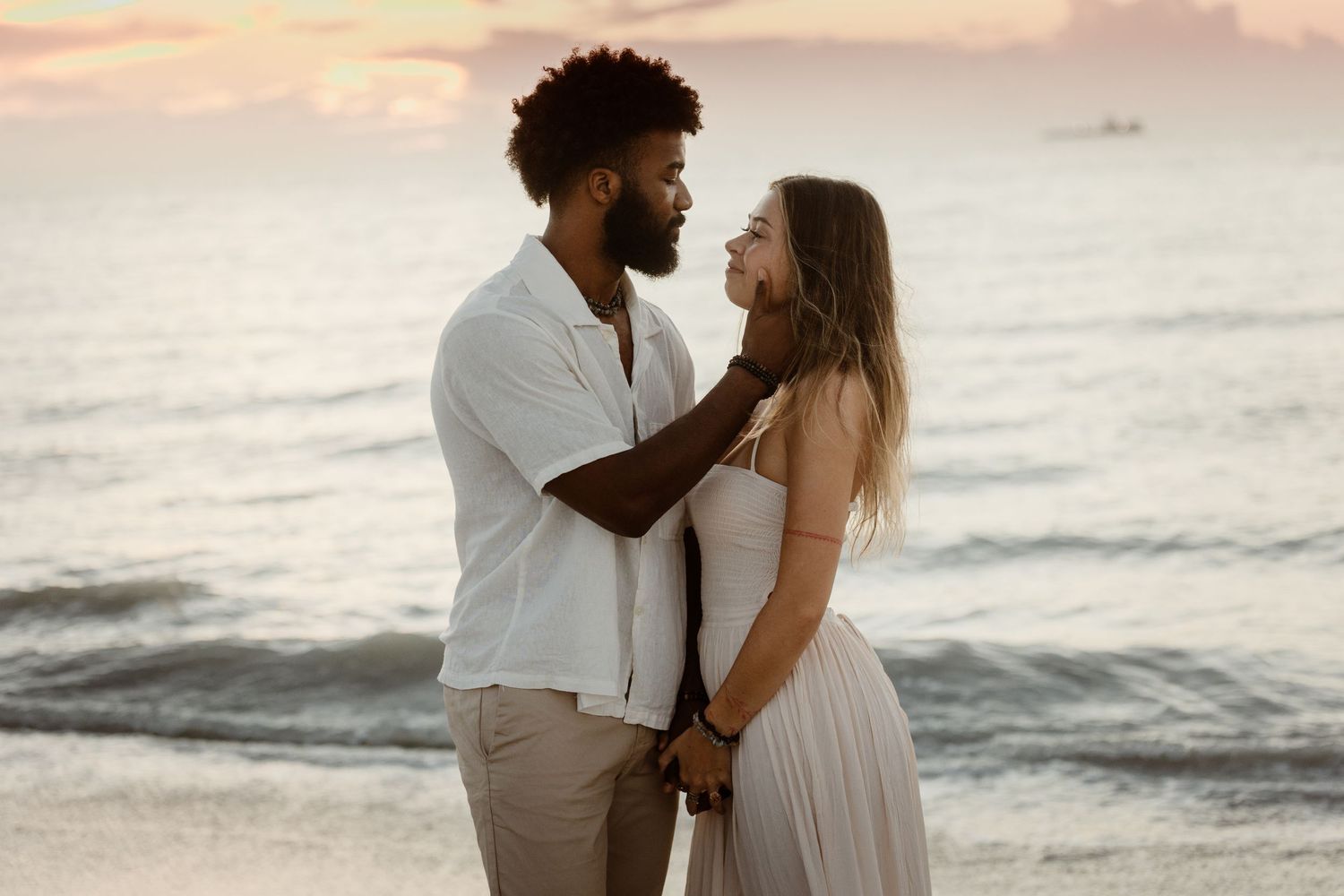 A couple shares a romantic moment on the beach at sunset with ocean waves in the background.