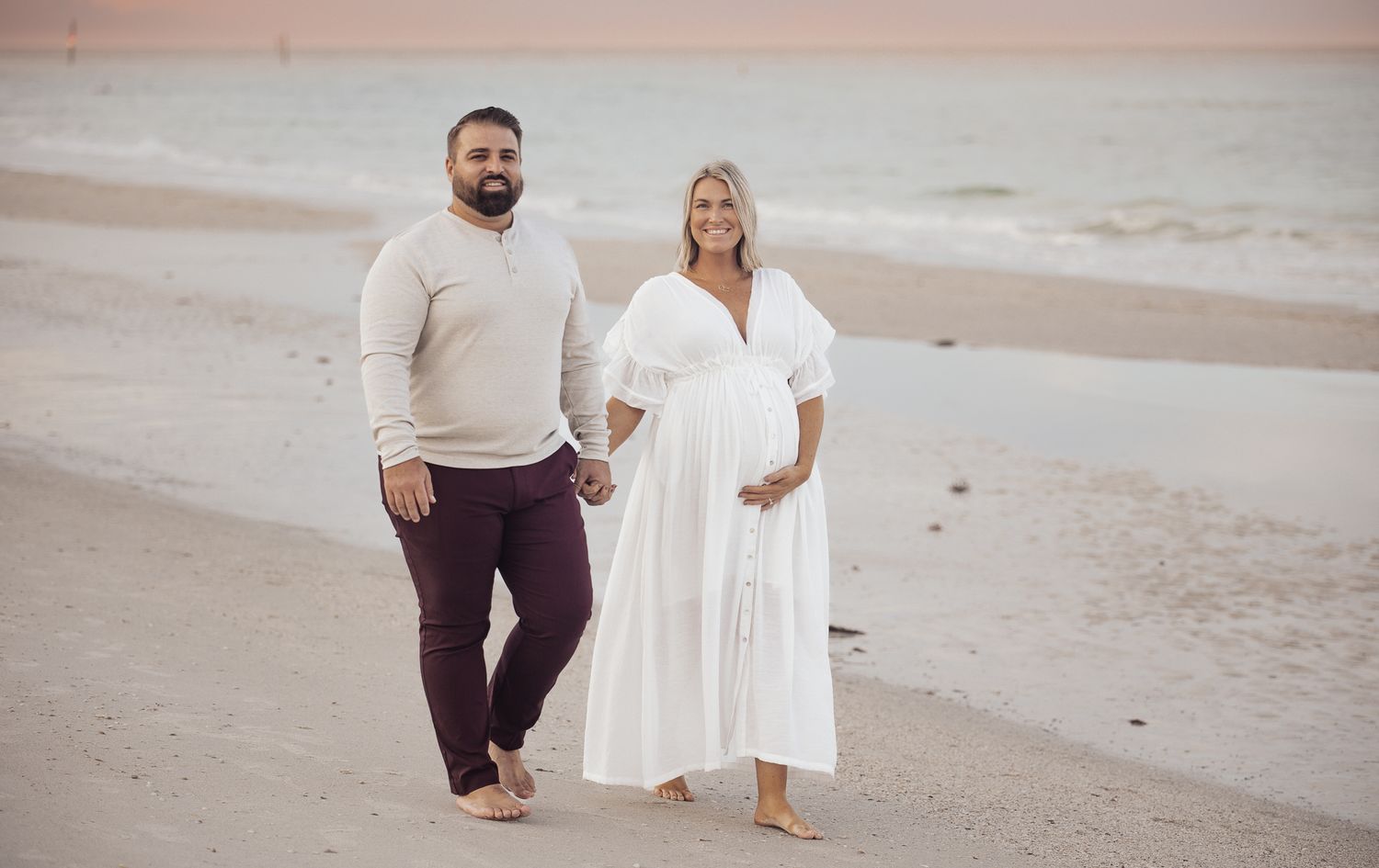A couple stands barefoot on a sandy beach at sunset wearing white and burgundy casual attire.