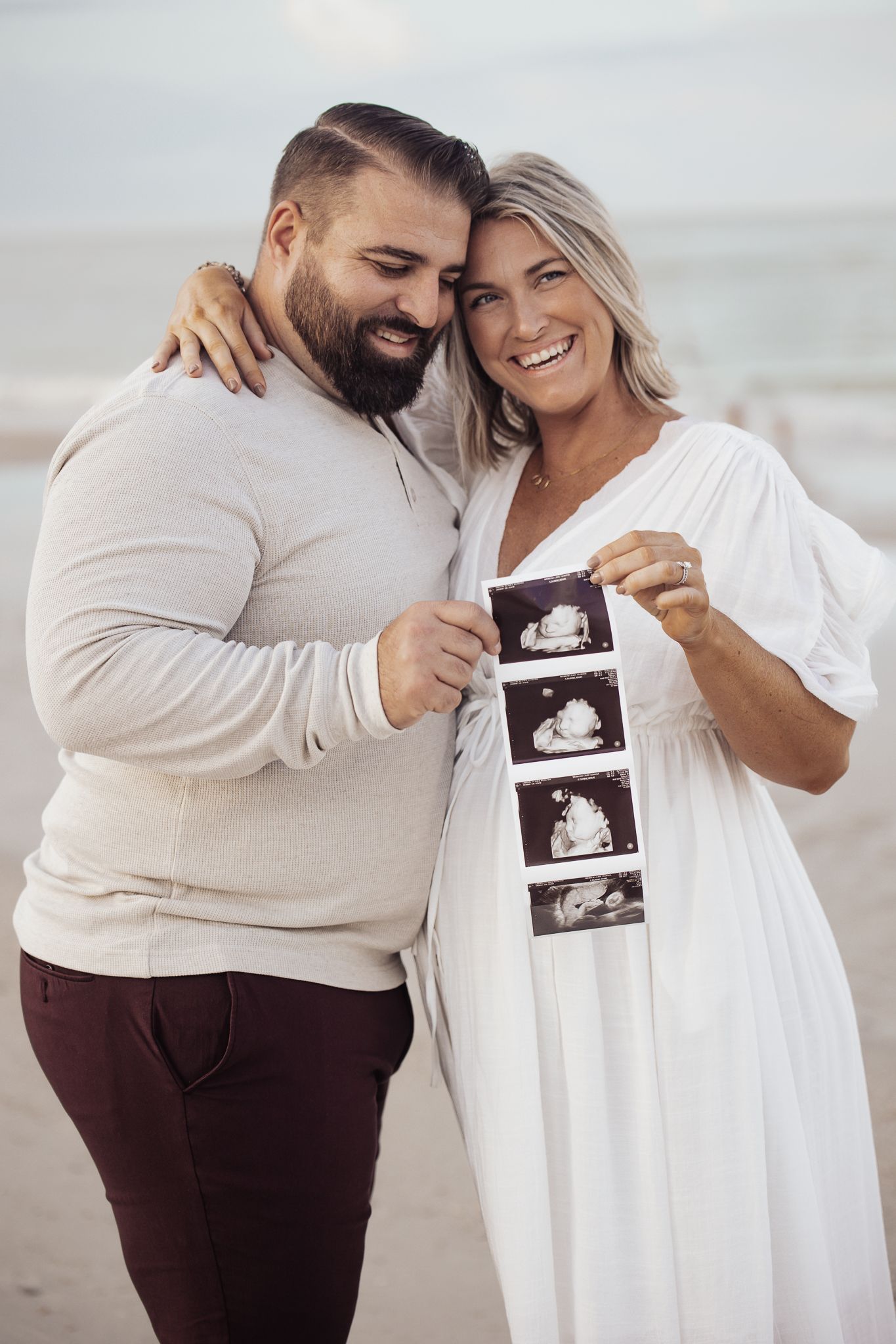 A couple on the beach shares a joyful moment while holding ultrasound photos in a pregnancy announcement photoshoot.