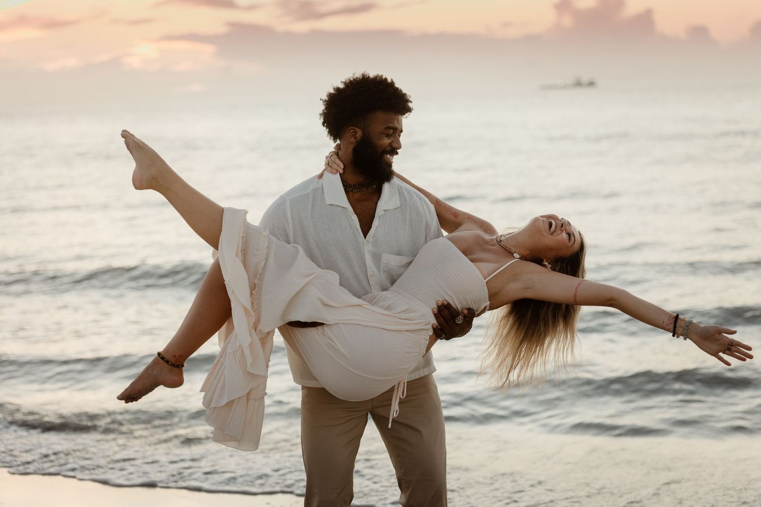 A couple in white clothing dances romantically together on a beach at sunset with ocean waves in the background.