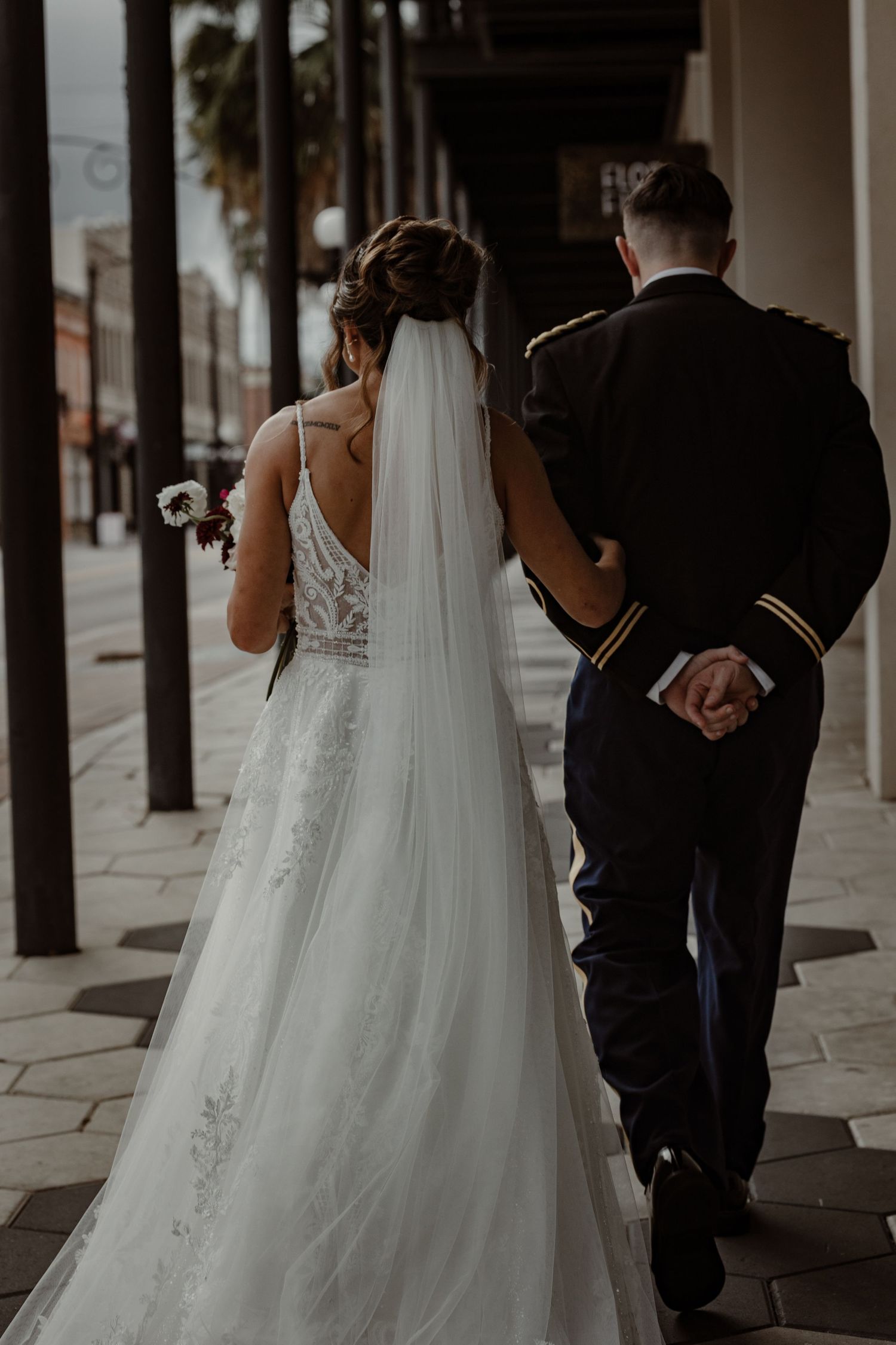 A bride in a flowing white lace gown walks along a covered sidewalk with wedding party members.
