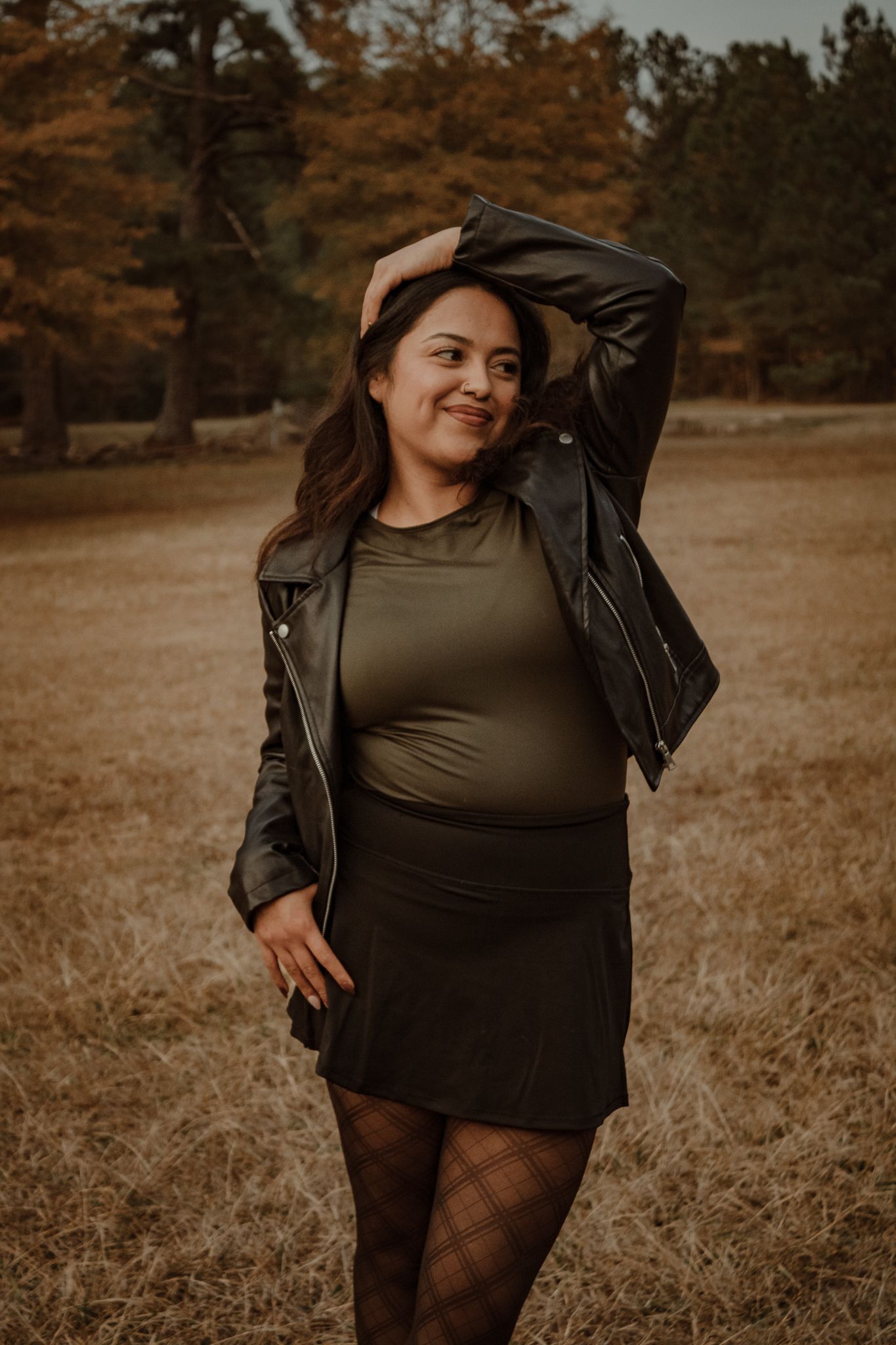 A person in a leather jacket and olive top poses in a field during autumn with warm moody lighting.
