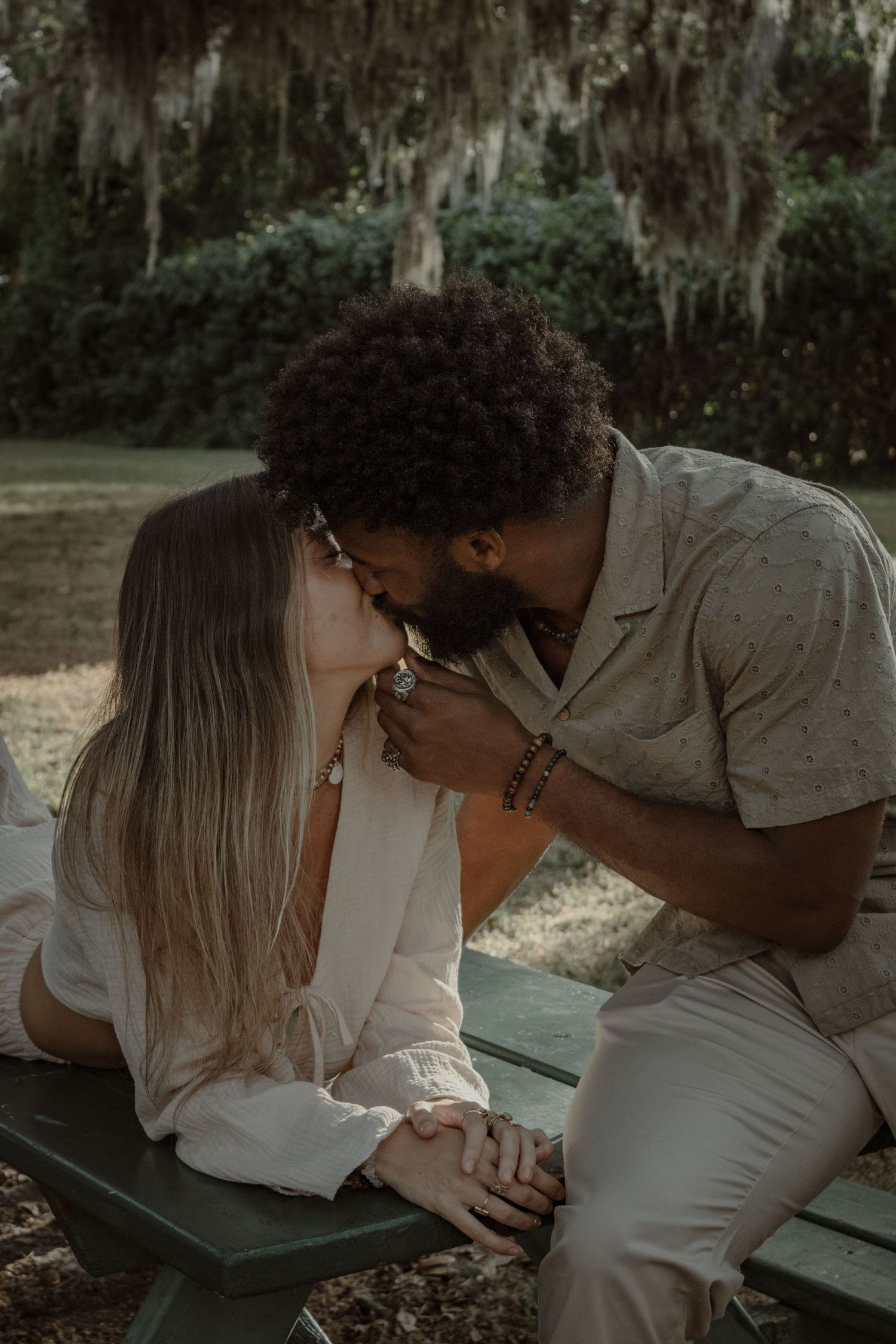 A romantic couple shares an intimate moment on a park bench under Spanish moss draped trees.