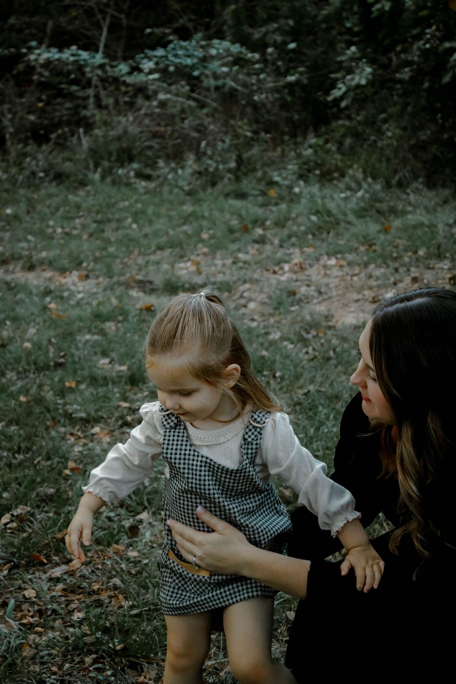 A small child in a gingham dress explores the outdoors while being supported by someone in black clothing.
