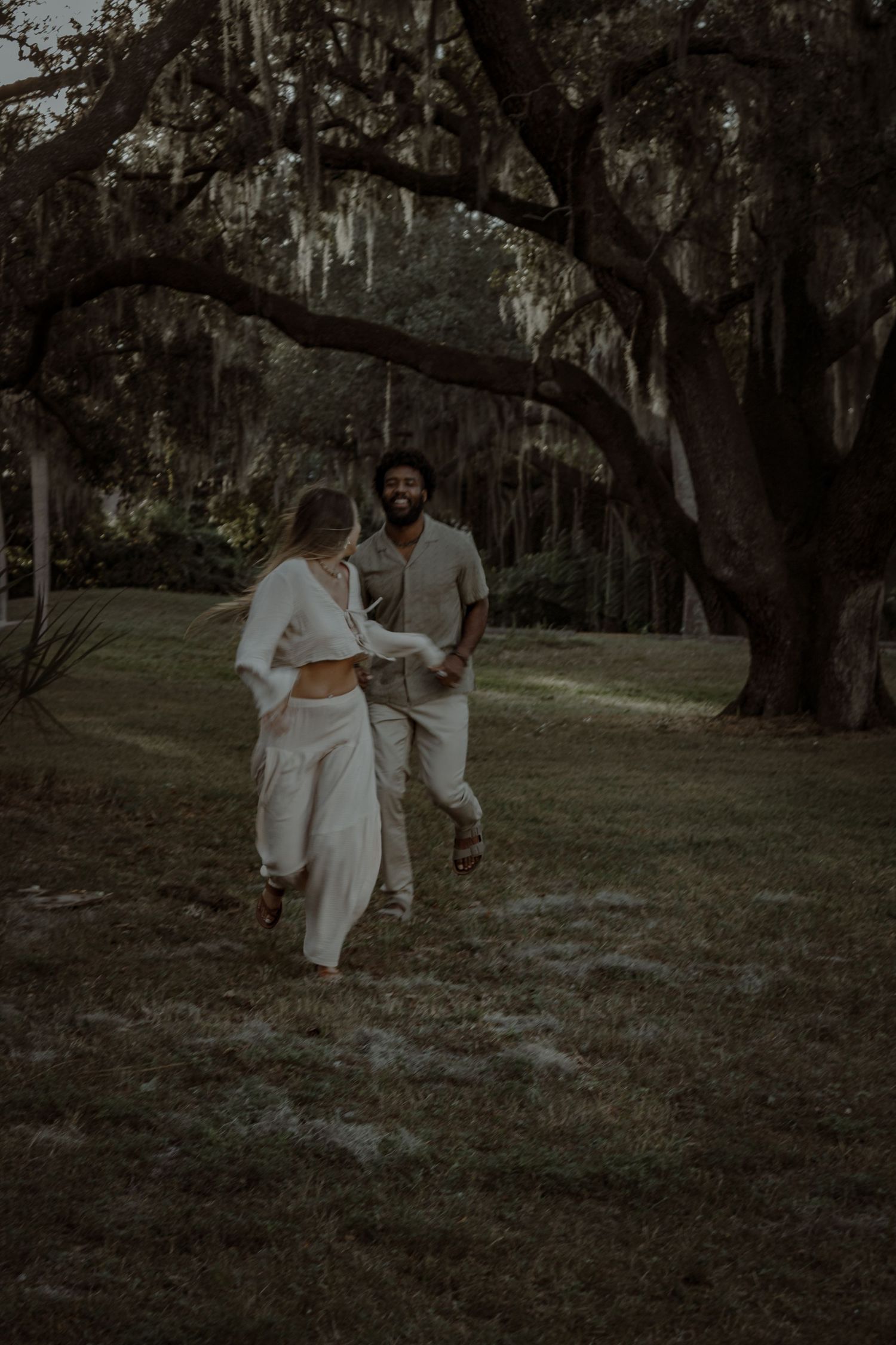 A couple dances together on grass beneath towering oak trees draped with Spanish moss.