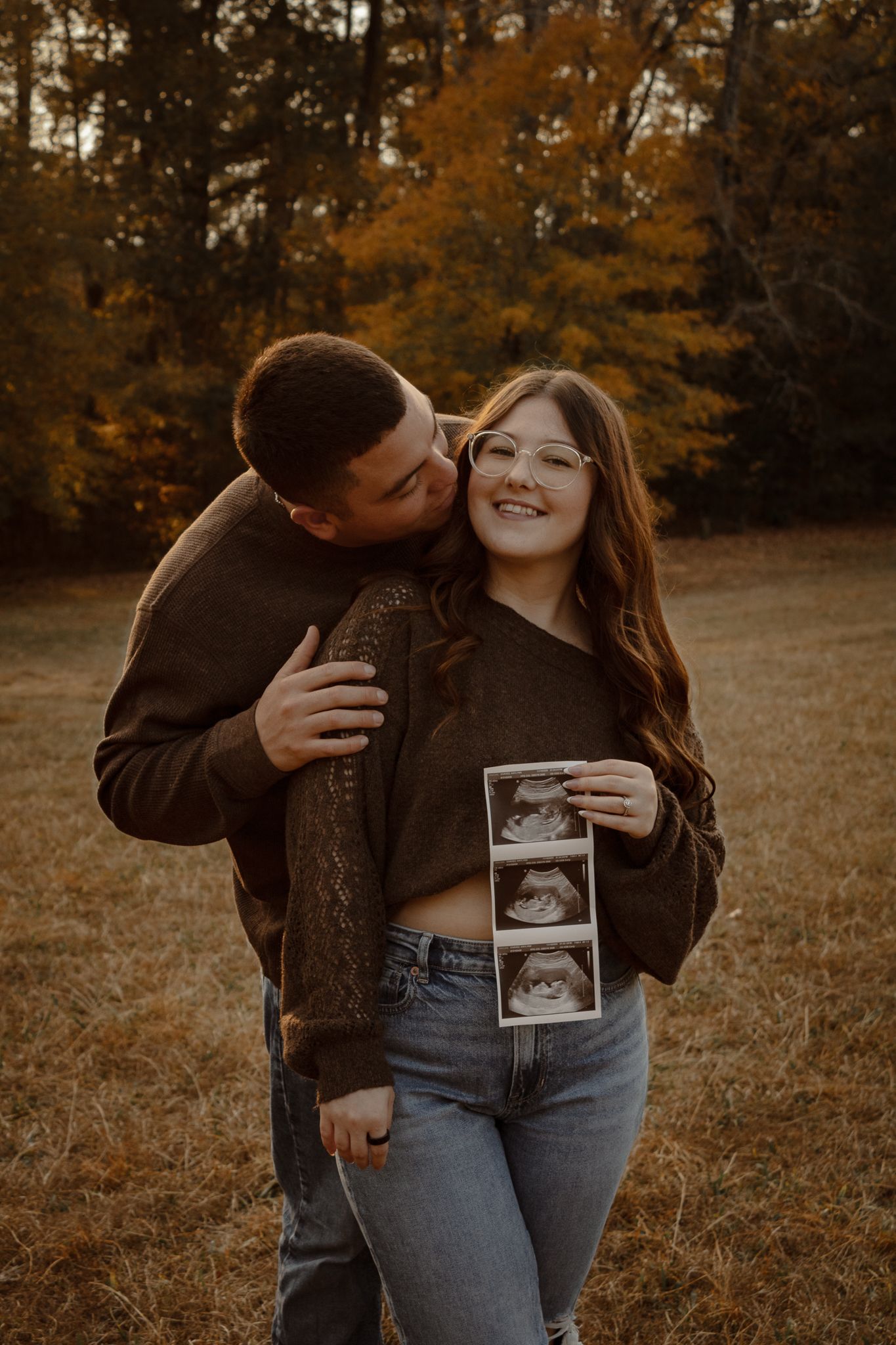 A couple embraces in a golden autumn field while holding an ultrasound photo during a pregnancy announcement photoshoot.