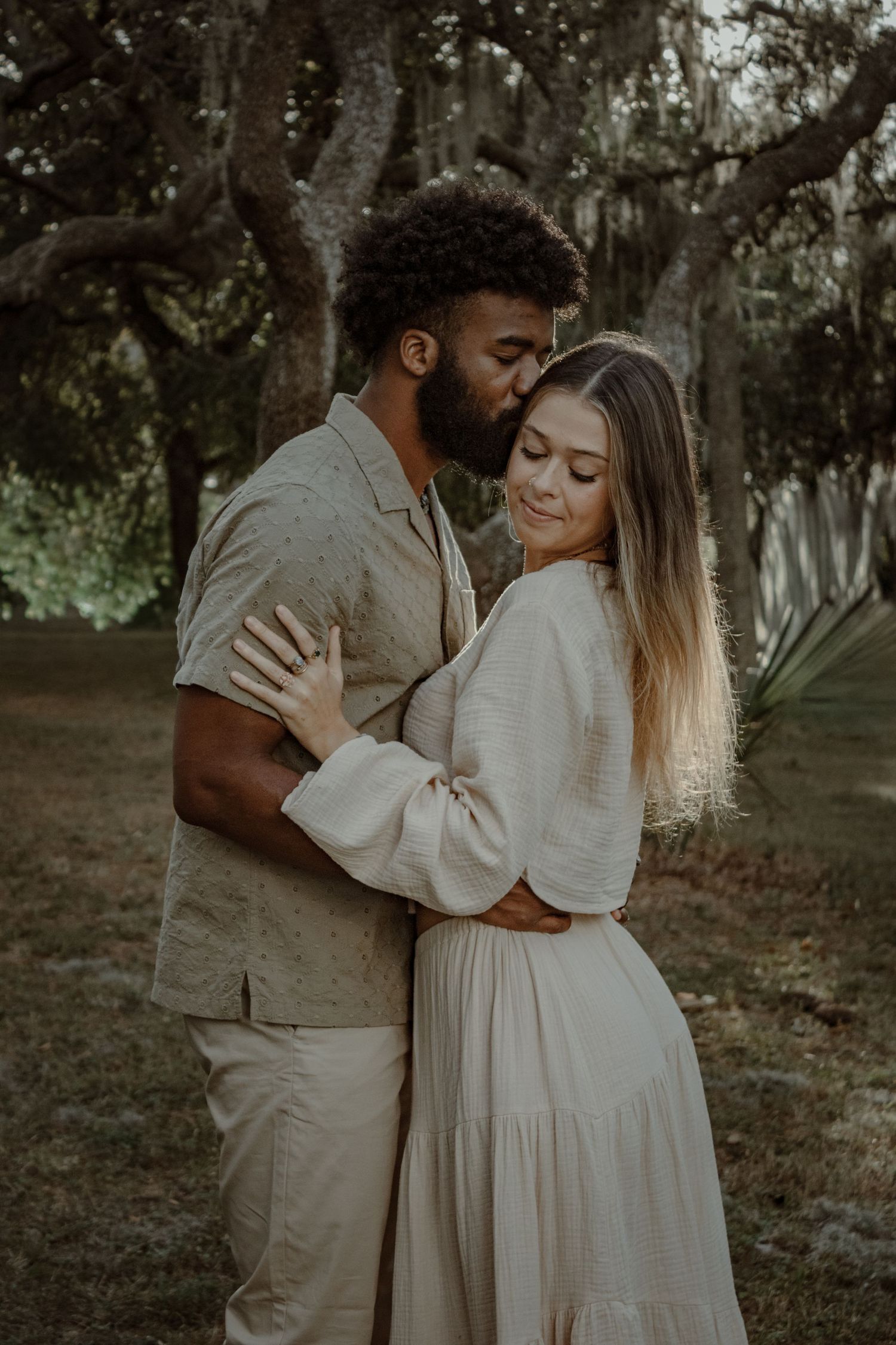 A romantic couple in white outfits embrace in a dreamy woodland setting with Spanish moss hanging from trees.