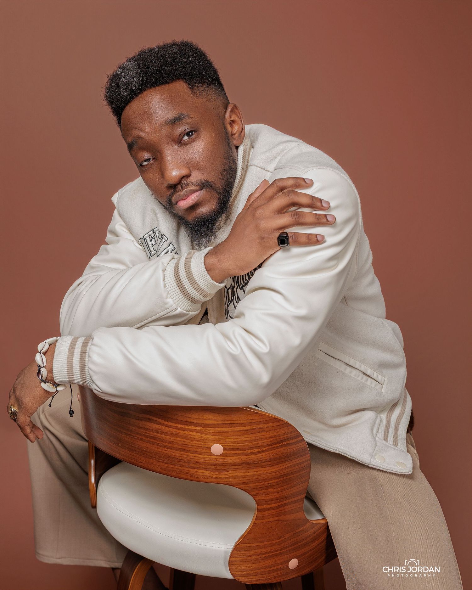 A person in a white sweater poses thoughtfully with a wooden chair against a brown studio backdrop in a series of portraits.