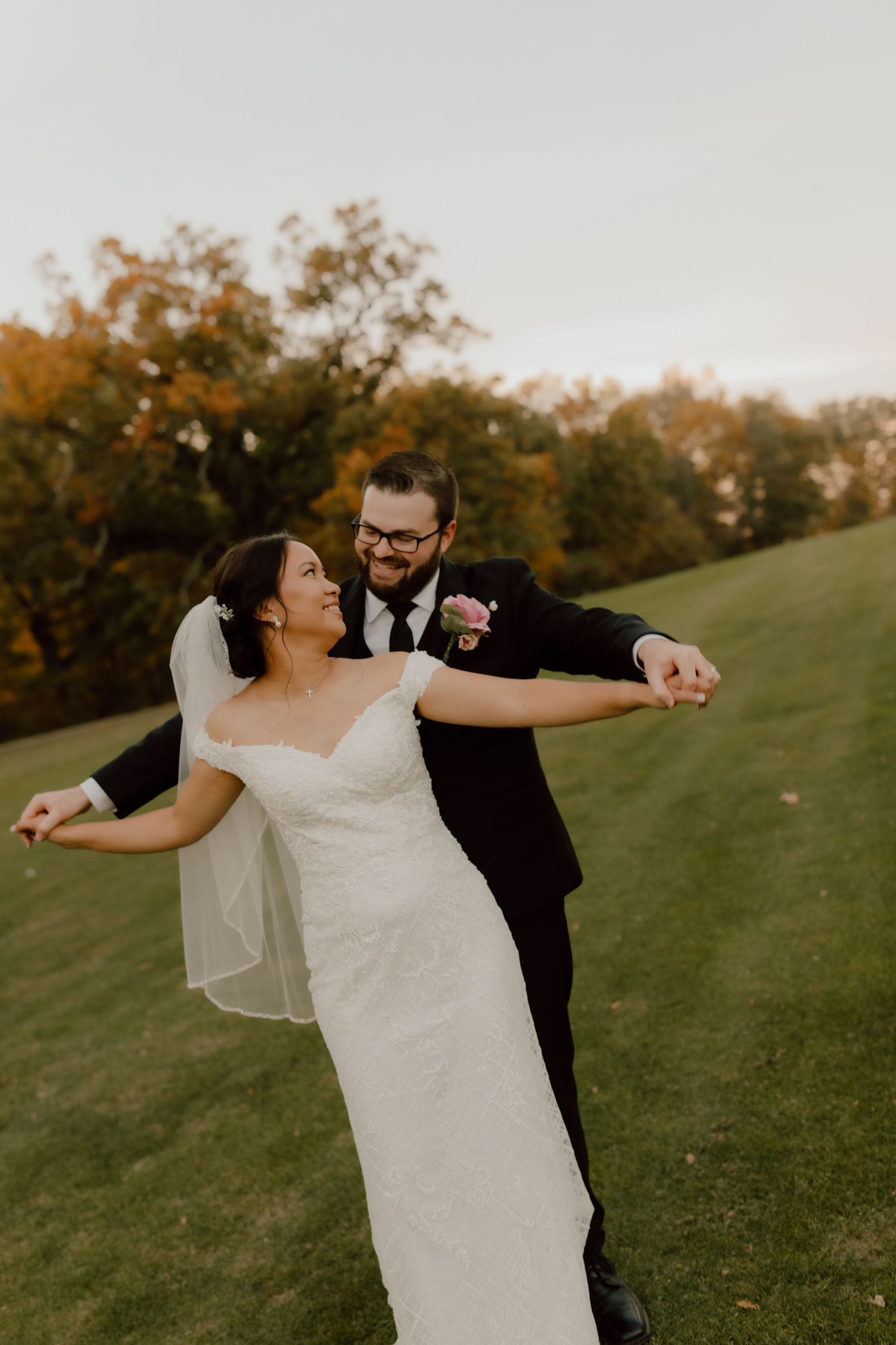 A joyful bride and groom dance outdoors on a green lawn surrounded by autumn trees on their wedding day.