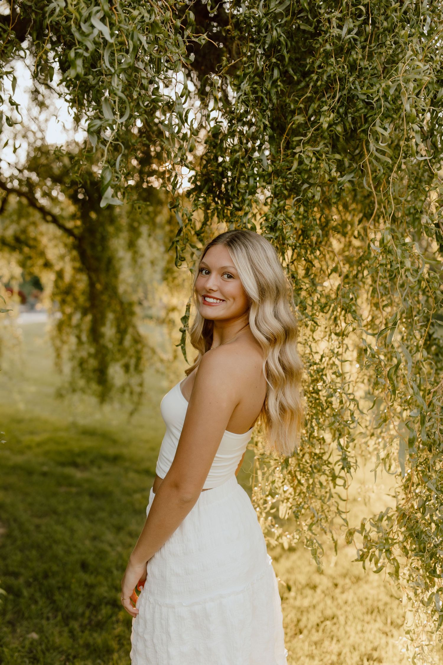 Dreamy golden hour portrait beneath weeping willow branches in white summer dress.