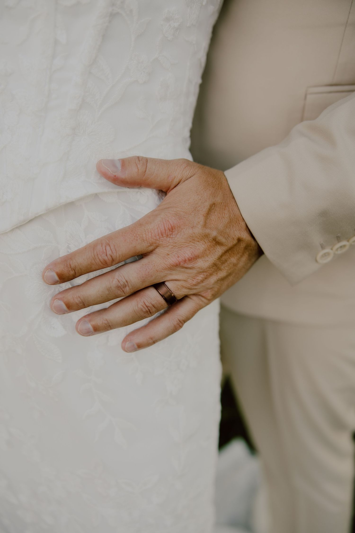 Close-up of grooms hand with black wedding band resting on brides white lace wedding dress.
