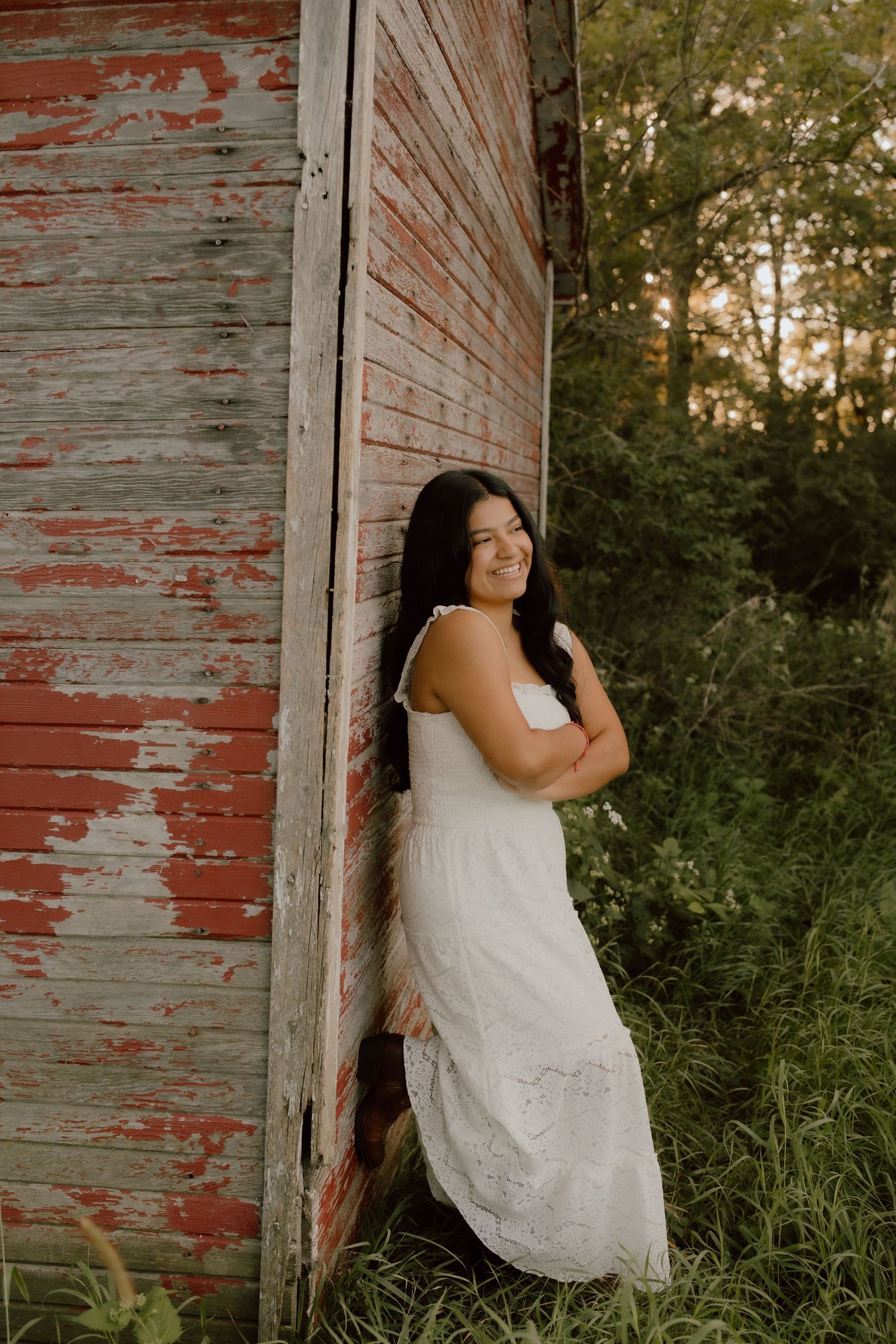 Person in white lace dress posing against rustic red barn wall at sunset.