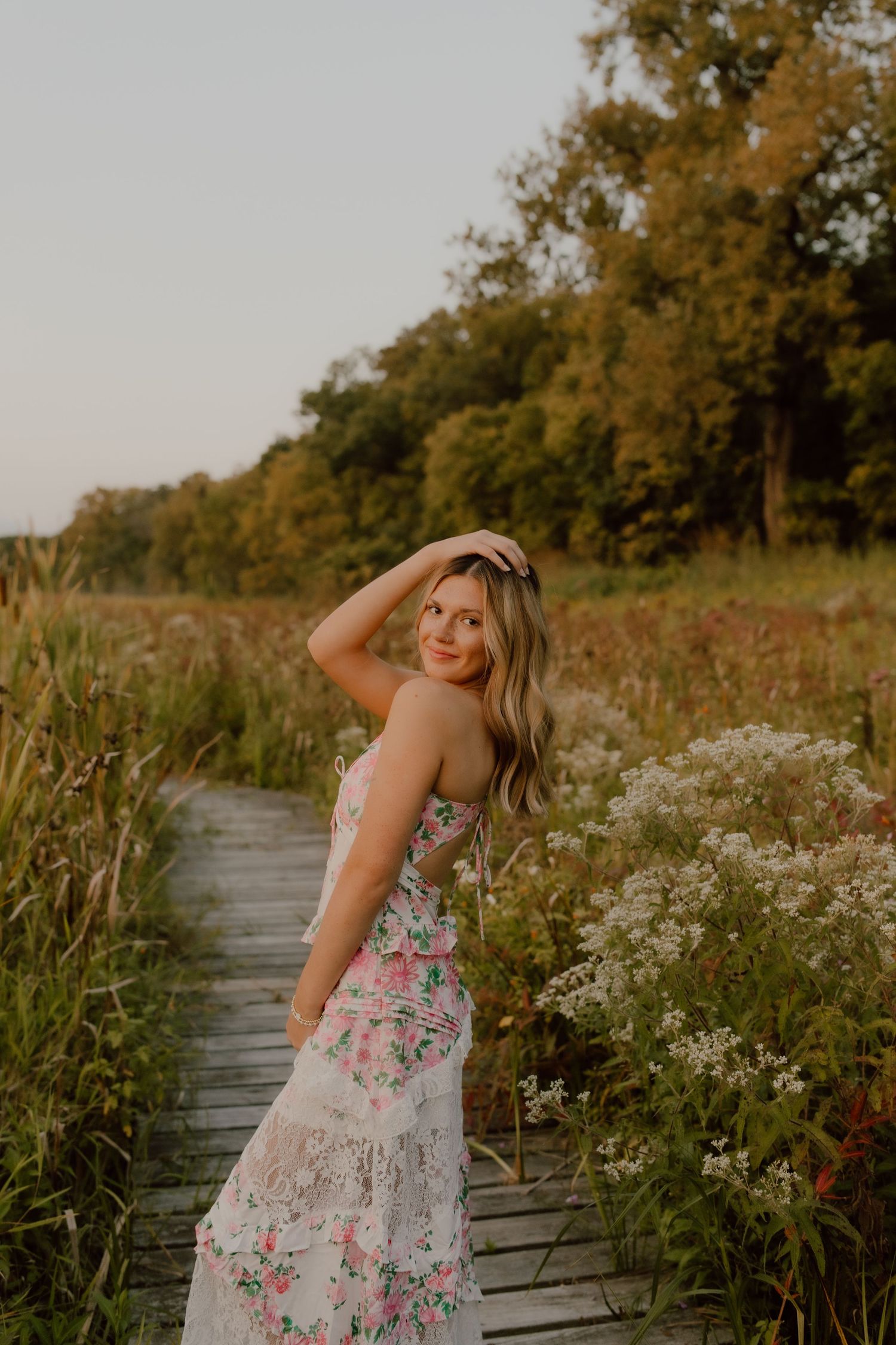A floral dress-clad figure poses on a wooden boardwalk surrounded by wildflowers at sunset.