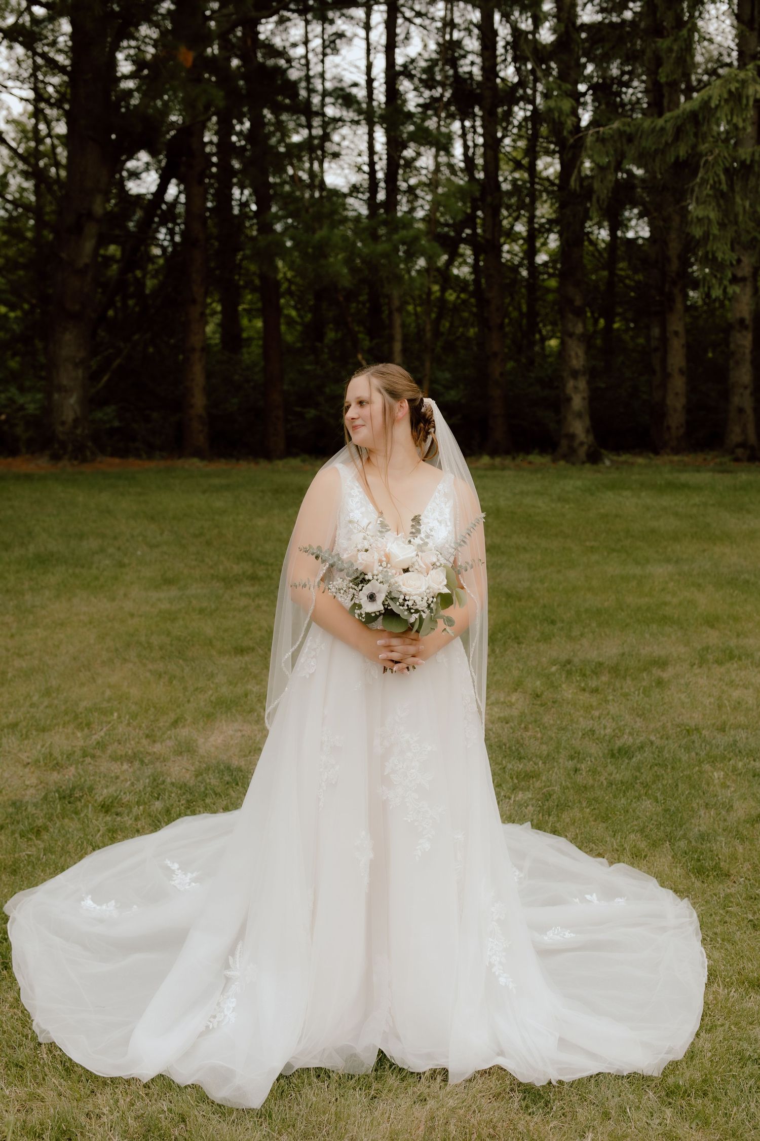 Bride in elegant white lace gown and veil holds bouquet while standing on green lawn near trees.