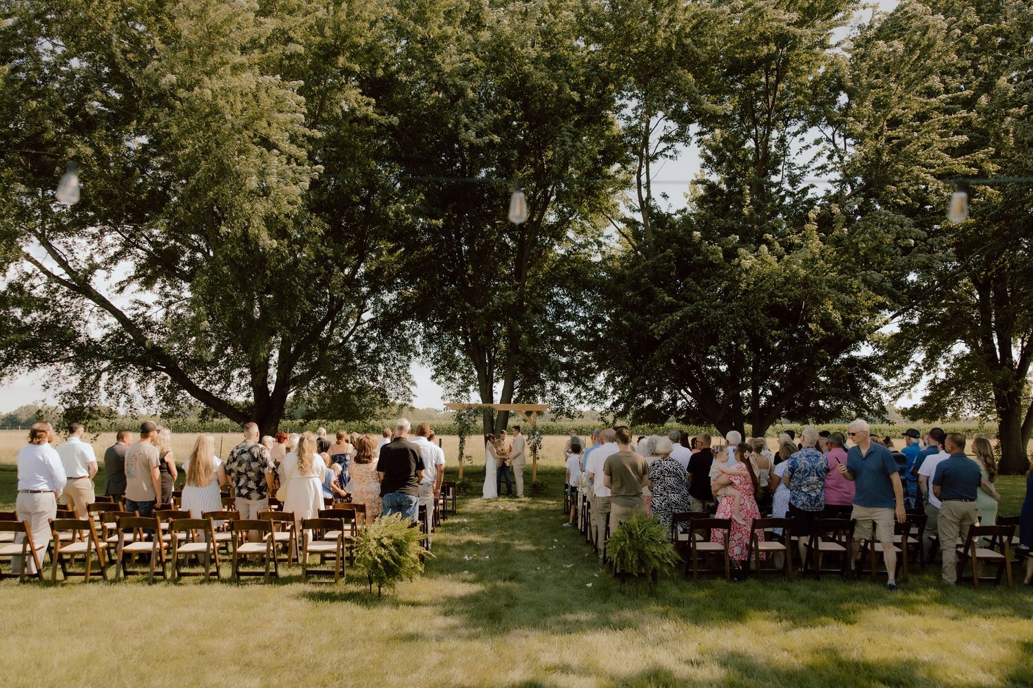 Outdoor wedding ceremony under large trees with guests seated on wooden chairs in a lush green garden setting.