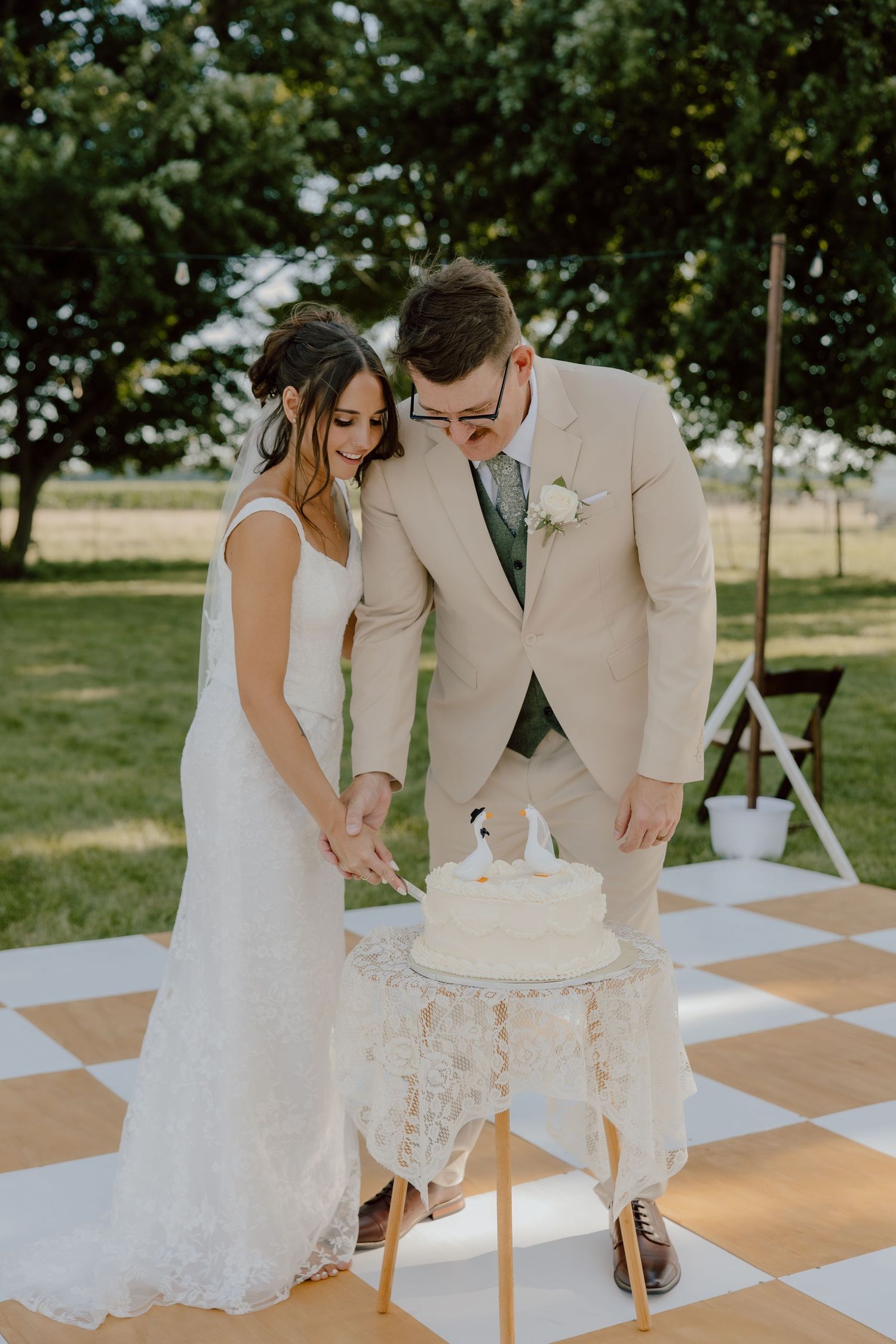 A bride and groom cut their white wedding cake outdoors on a checkered dance floor surrounded by lush greenery.