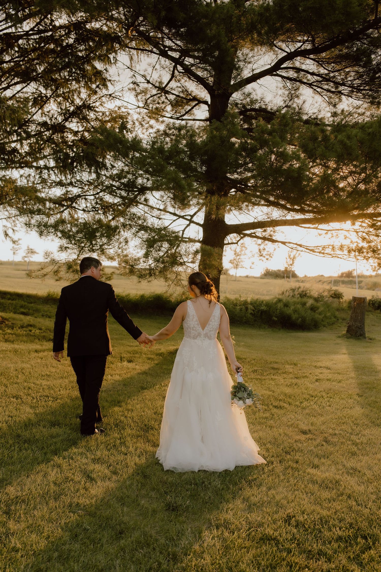 Bride and groom hold hands walking through golden sunset light on a green lawn near a large tree.