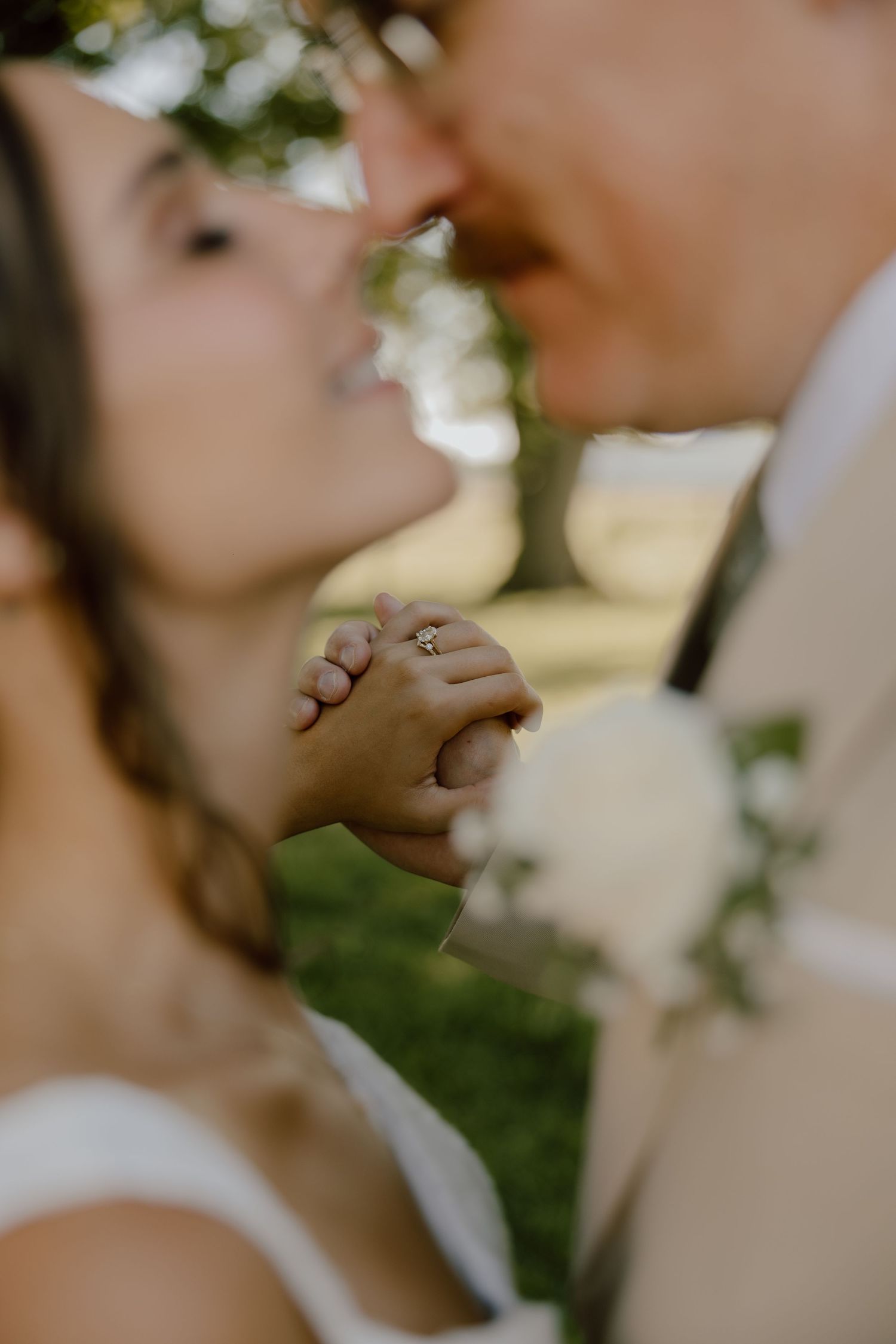 Close-up of bride and groom kissing while holding hands, showcasing her diamond engagement ring outdoors.