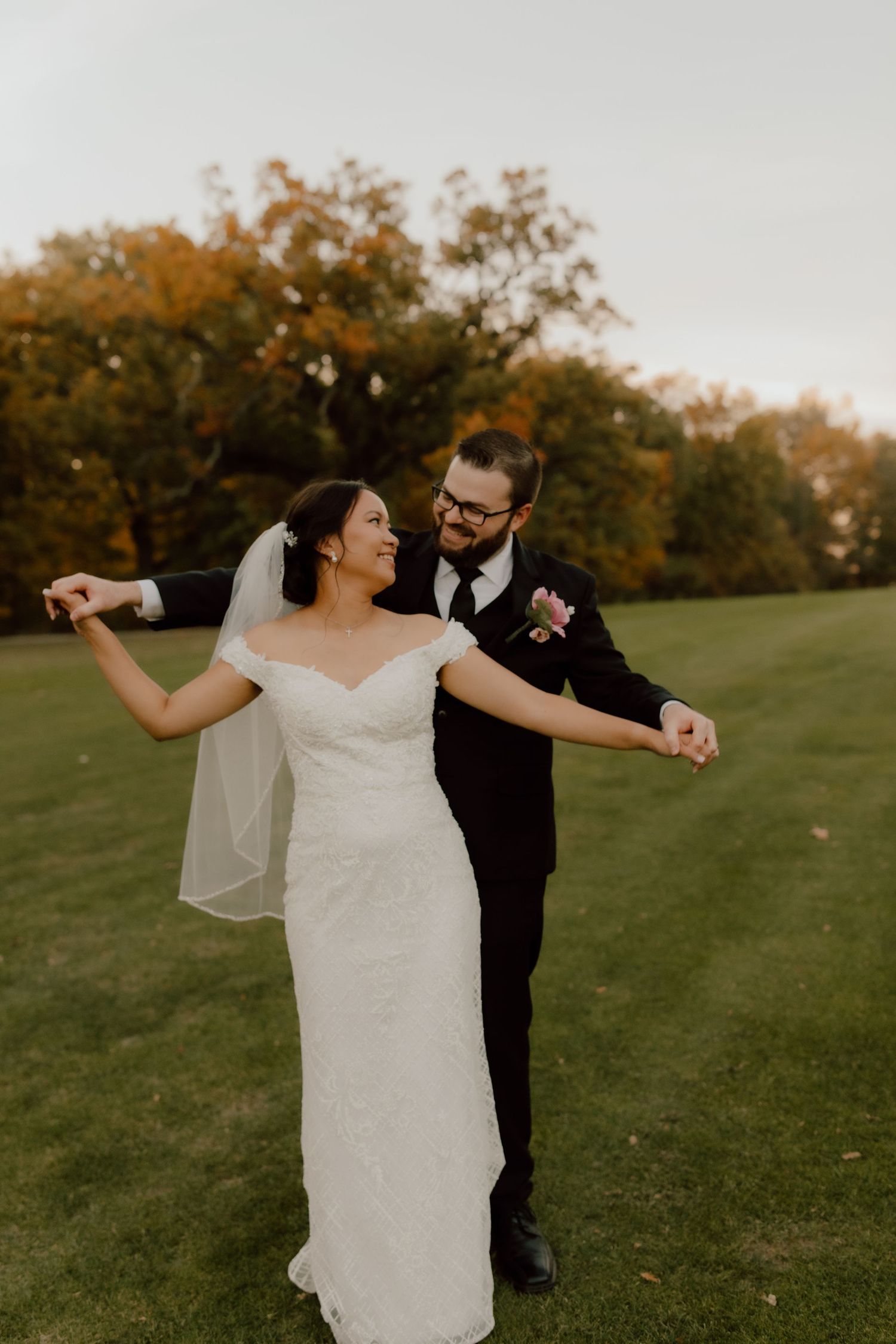 A bride and groom dance joyfully on a green lawn surrounded by autumn trees, she in a white gown and he in a black suit.