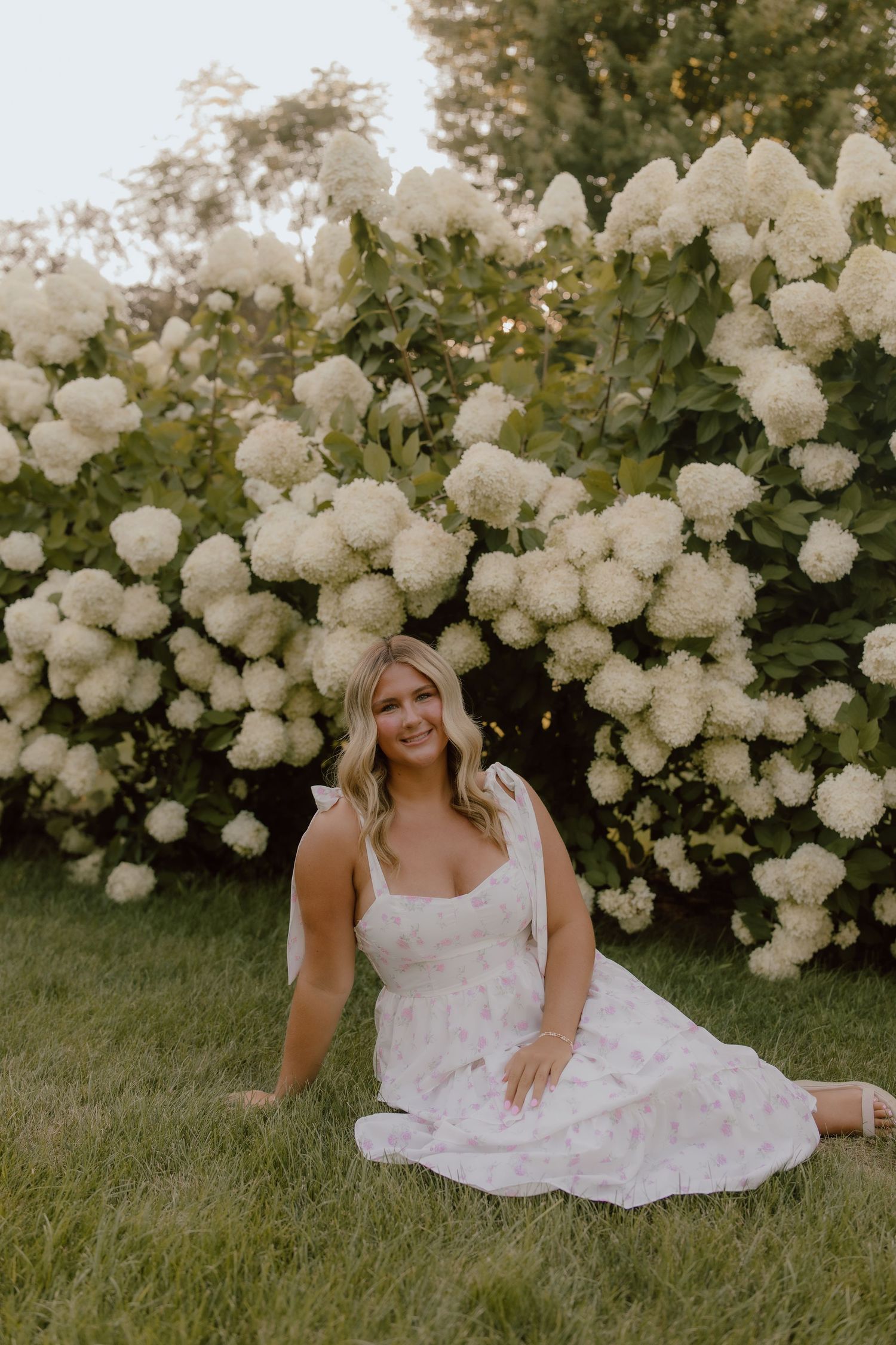 A person in a white sundress sits gracefully among blooming white hydrangea bushes in a garden setting.