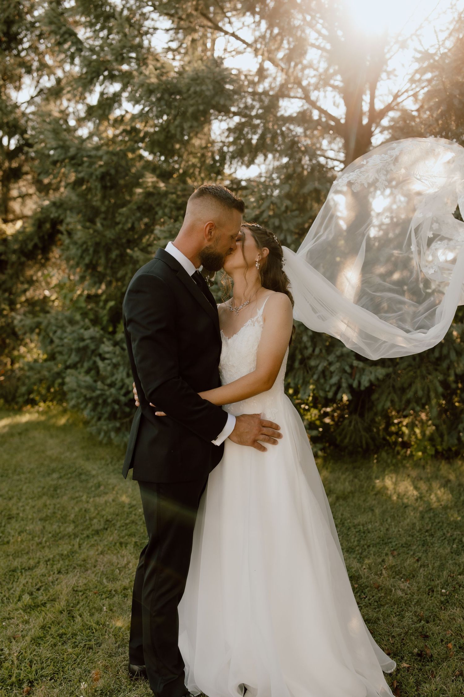 A romantic wedding kiss captured in golden sunlight as the bride's veil flows dramatically.