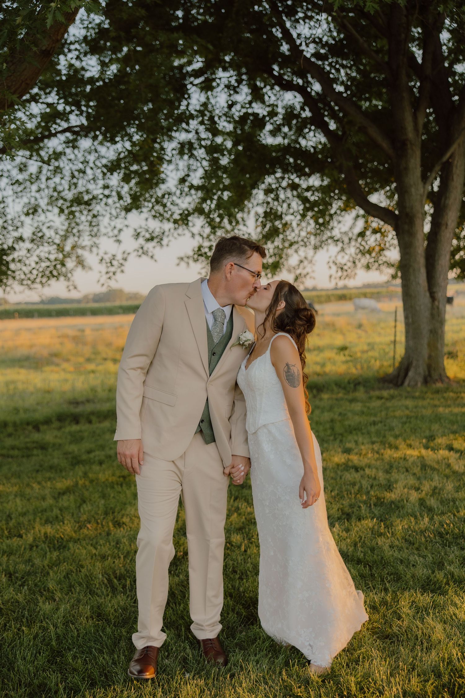 A couple shares a kiss in a sunlit field, the groom in a tan suit and bride in a white gown, holding hands under a large tree.