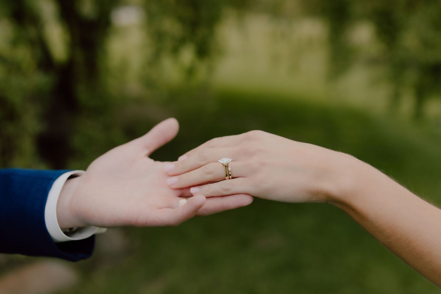 Close-up of a bride wearing a gold diamond engagement ring as she reaches toward the groom's hand outdoors.
