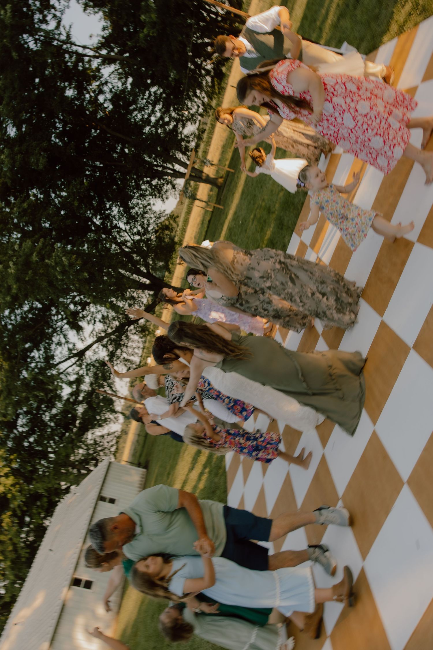Guests dancing on a checkered dance floor at an outdoor summer wedding reception surrounded by lush greenery.