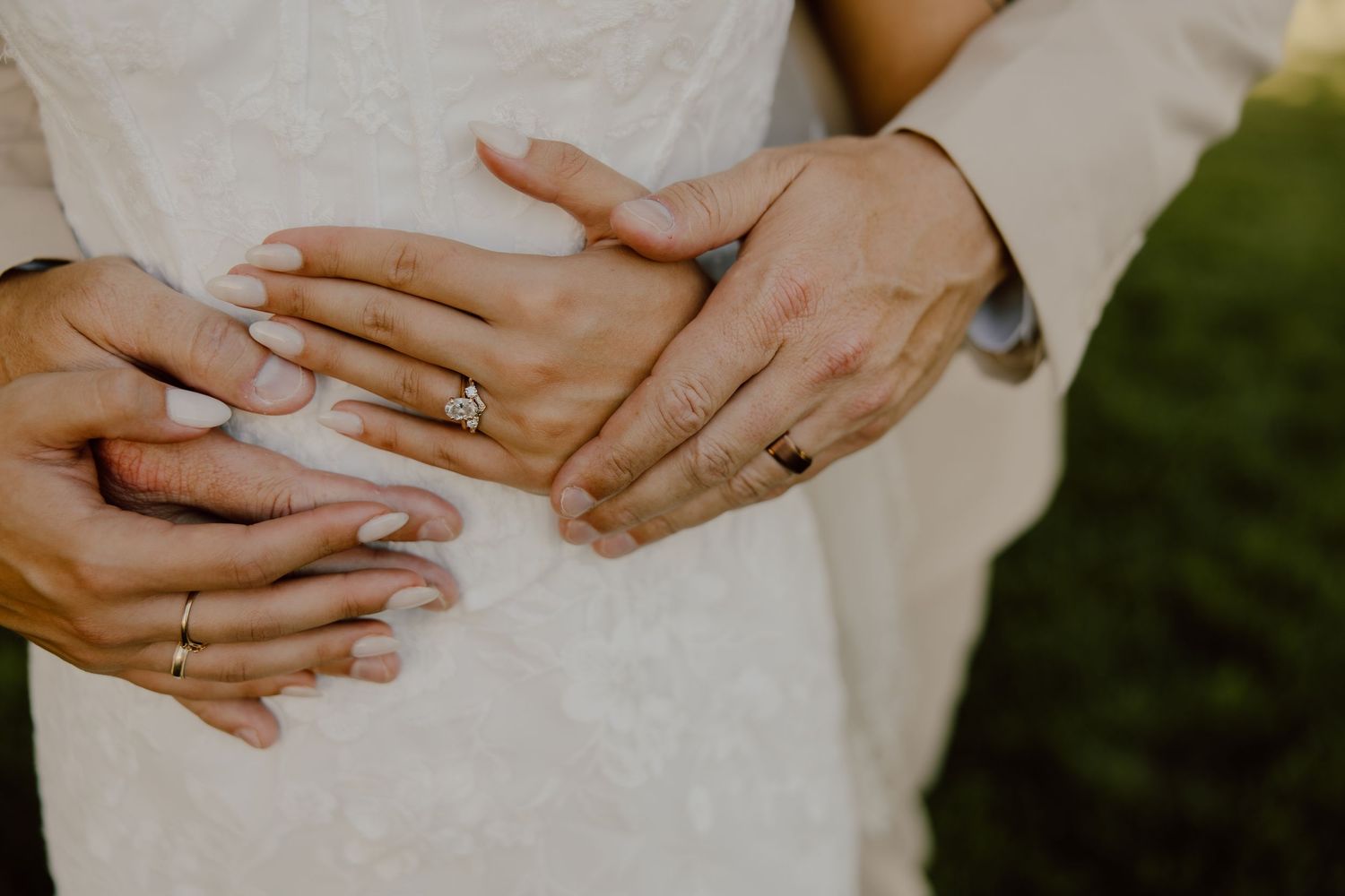 Close-up of bride and groom hands showing wedding rings, bride in white lace dress held by groom outdoors.