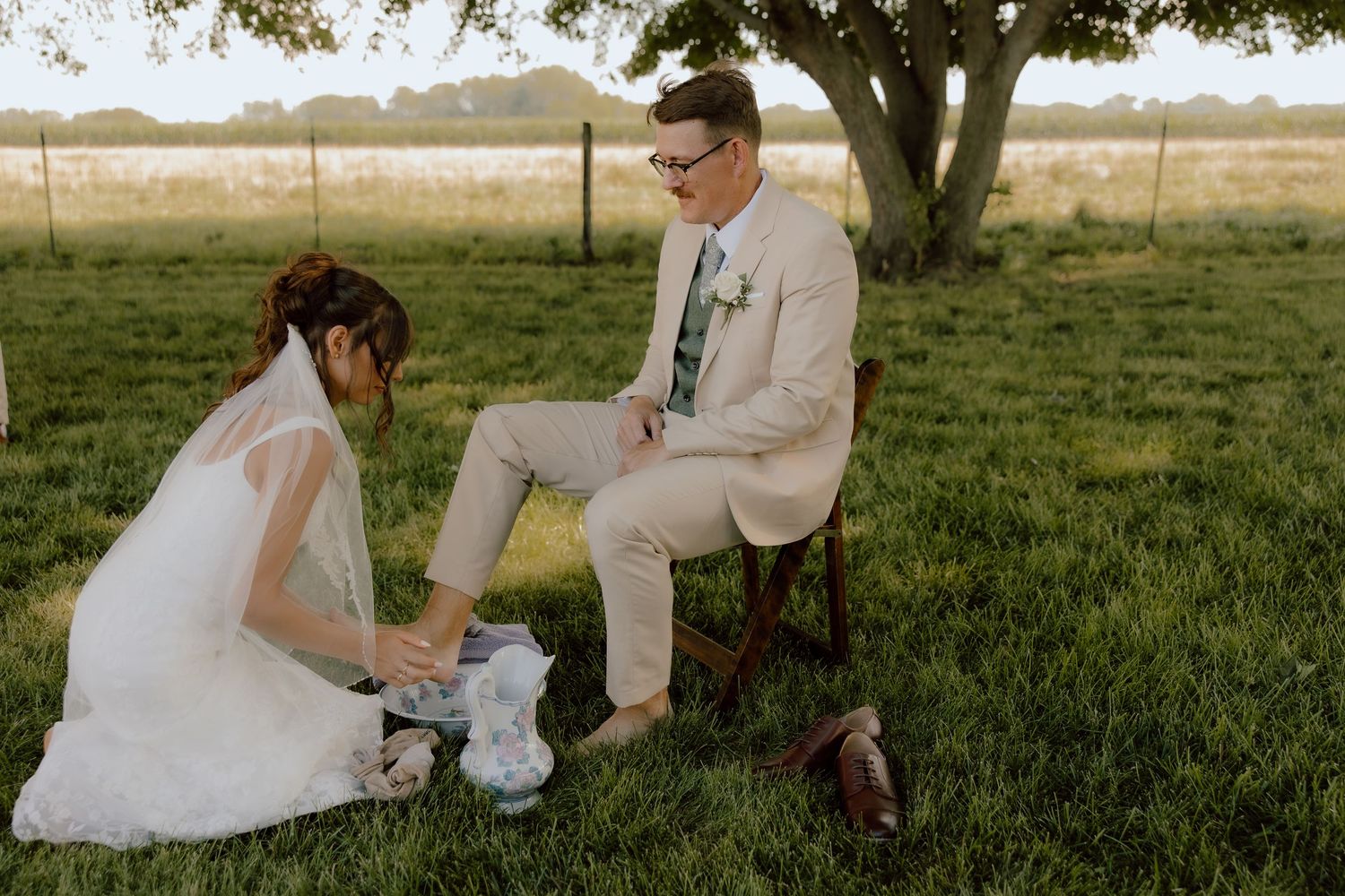 Bride kneeling on grass helping groom with shoes during outdoor wedding ceremony under large tree.